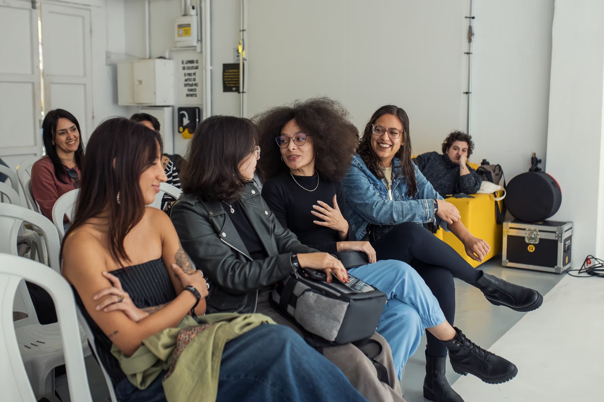 A group of young people sits on white chairs in a brightly lit room, chatting together.