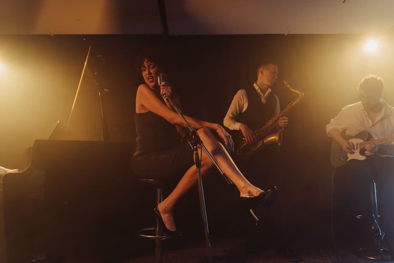 A female singer sits on a stool while two band members play instruments on stage.