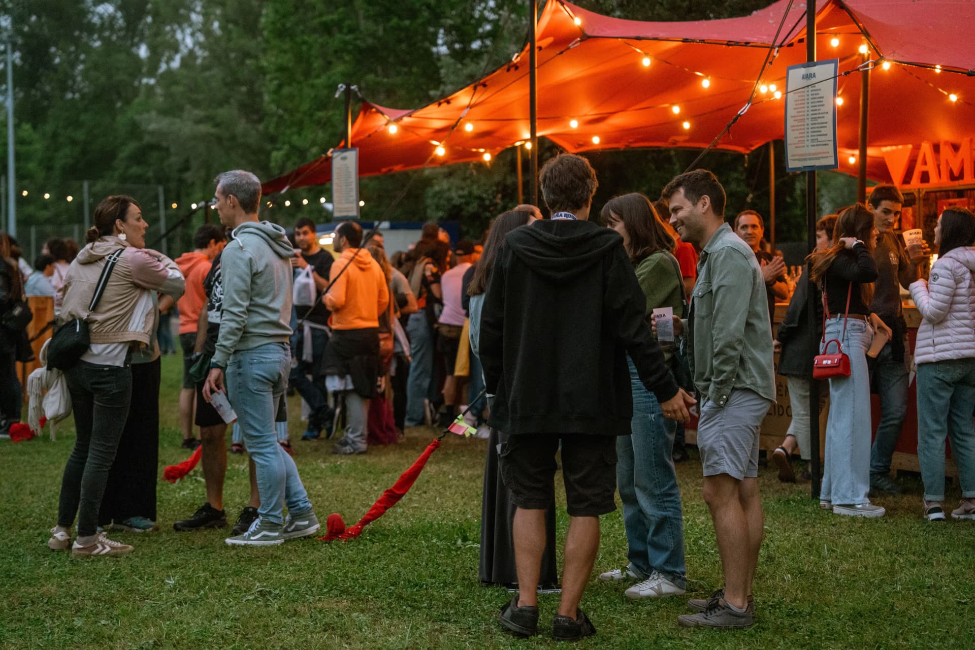 People socialize at an evening outdoor event under a large orange tent lit with warm string lights.