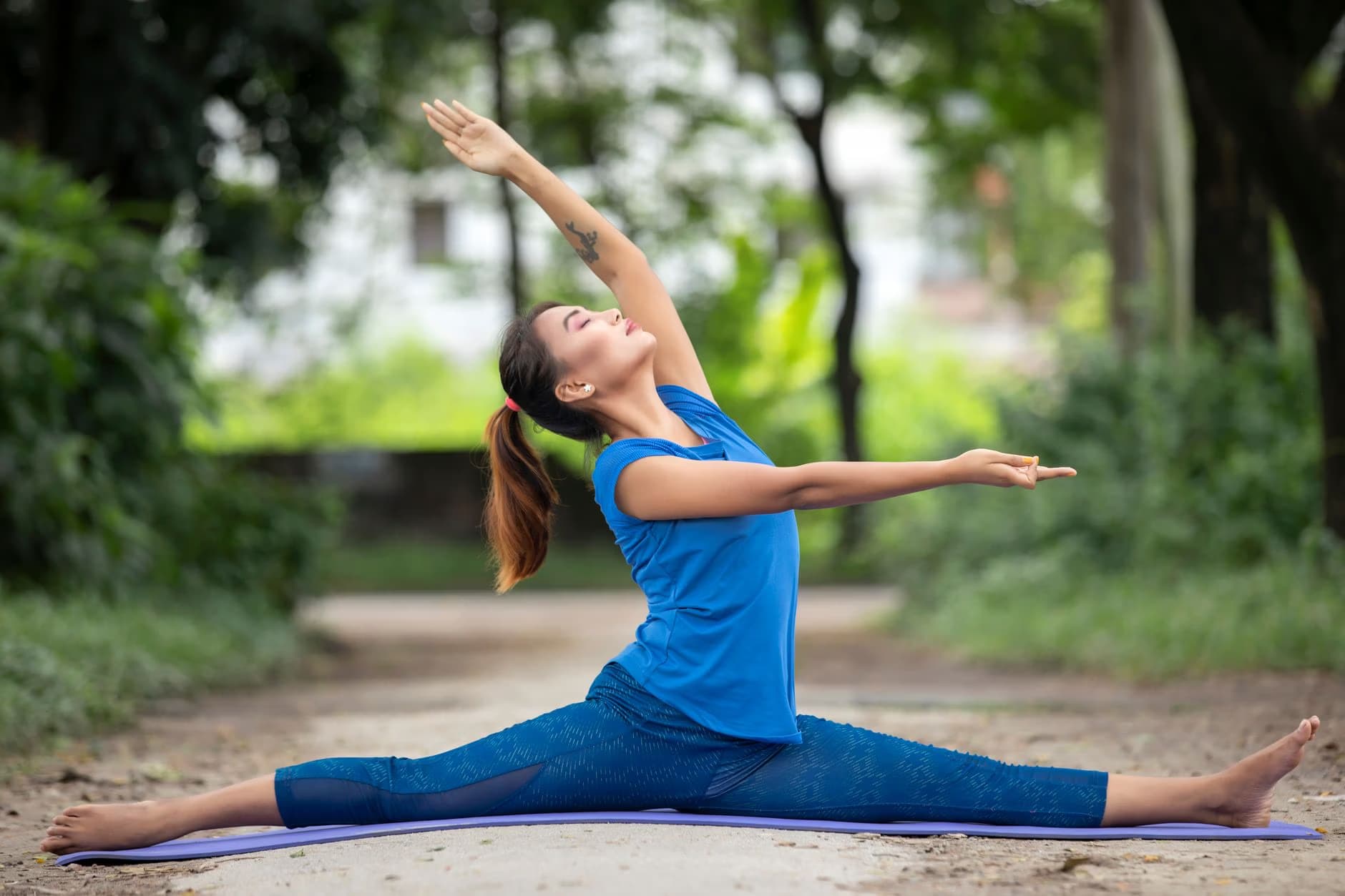 A woman in blue activewear performs a split yoga pose on a mat in a park.