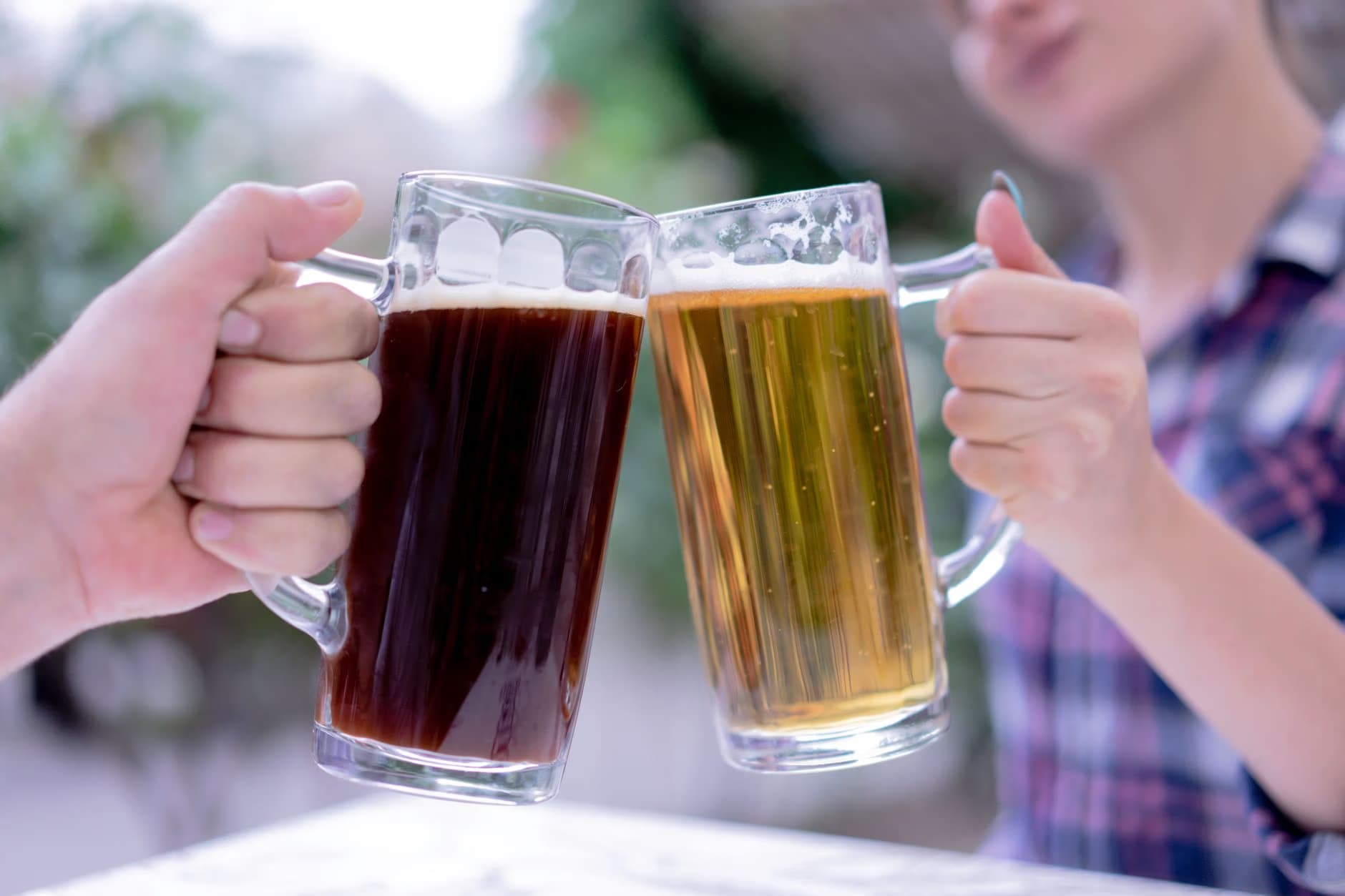 Two people clink glass mugs of dark and light beer in a cheers gesture.