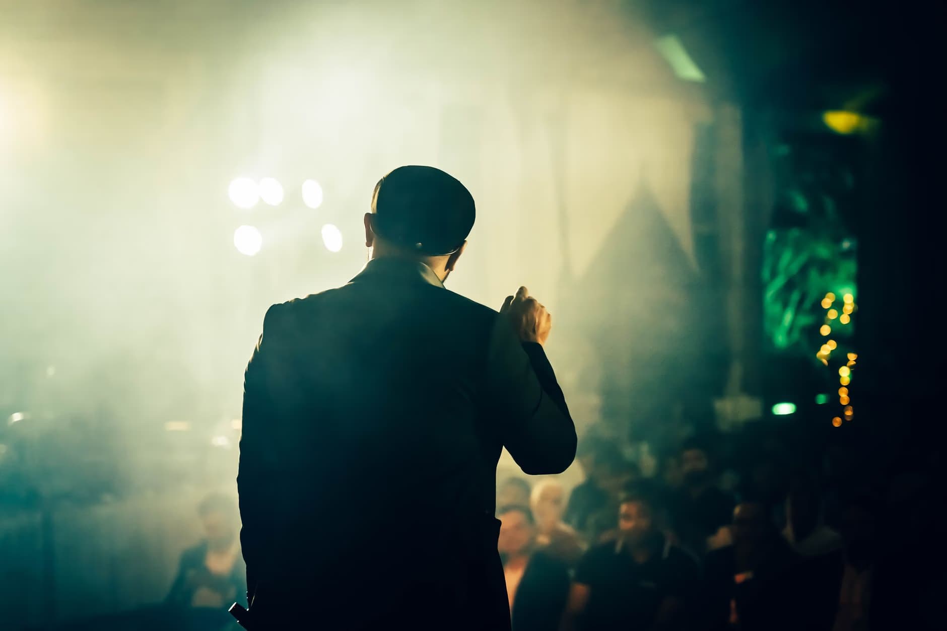 Mężczyzna w garniturze stoi na scenie, zwracając się do publiczności w słabo oświetlonej sali.