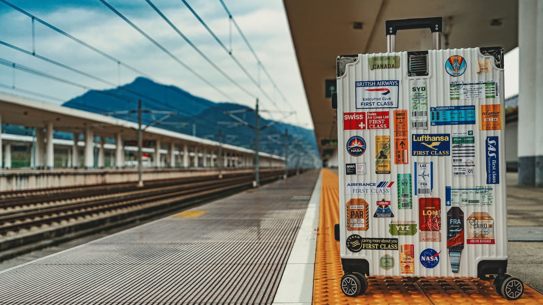 A suitcase covered in travel stickers stands on a train station platform before mountain scenery.
