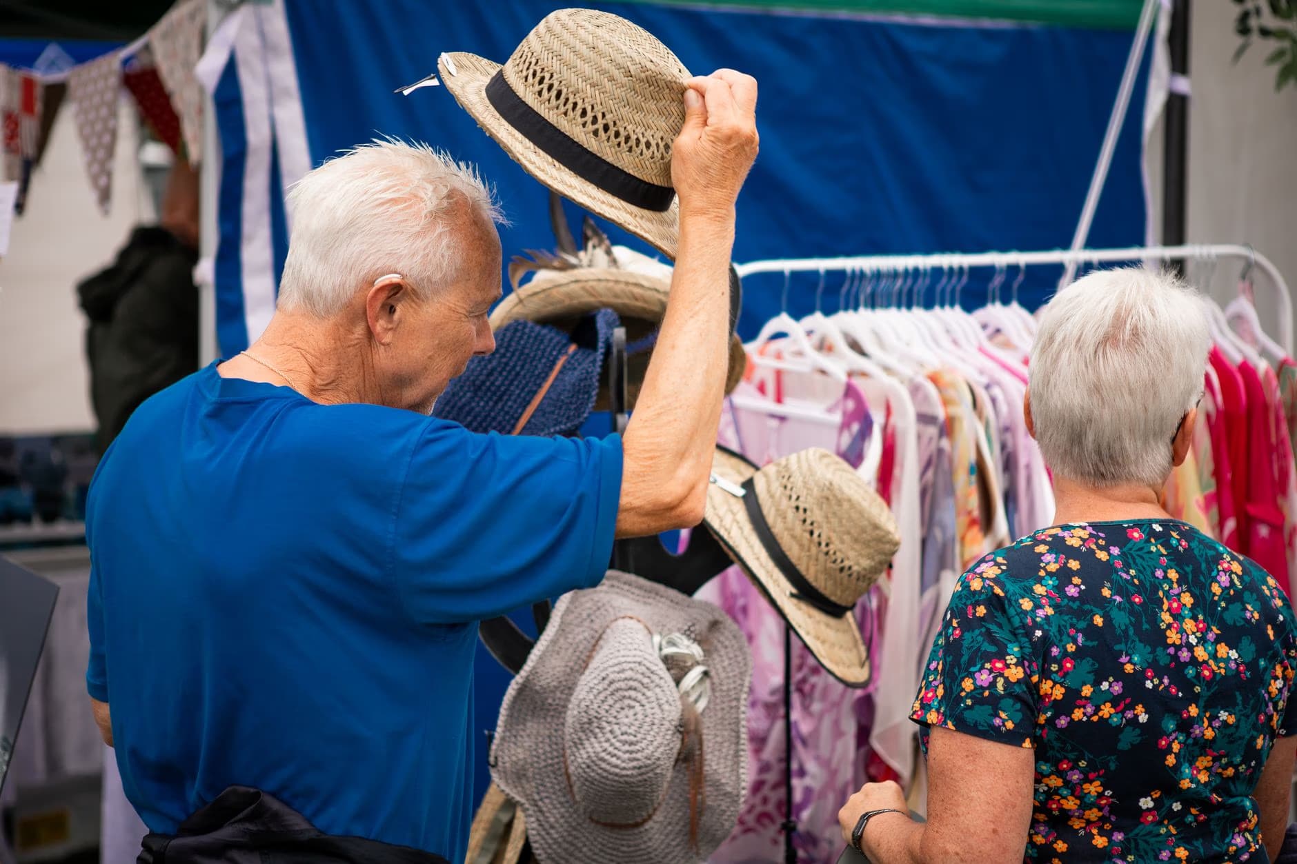 Ein älteres Paar betrachtet Hüte und Kleidung an einem Marktstand im Freien.