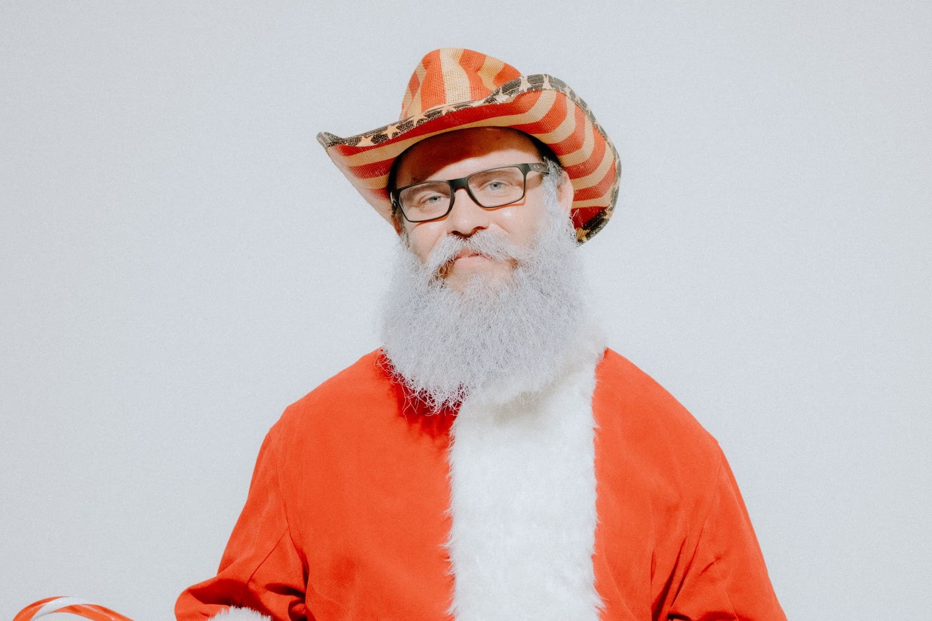 A man in a Santa Claus costume and a star-spangled cowboy hat smiles against a white background.