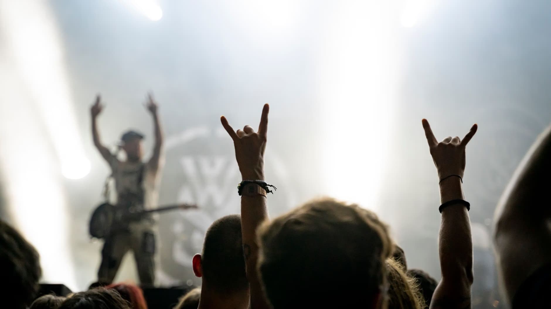 Concert crowd raising hands with rock signs in front of bright stage lights.