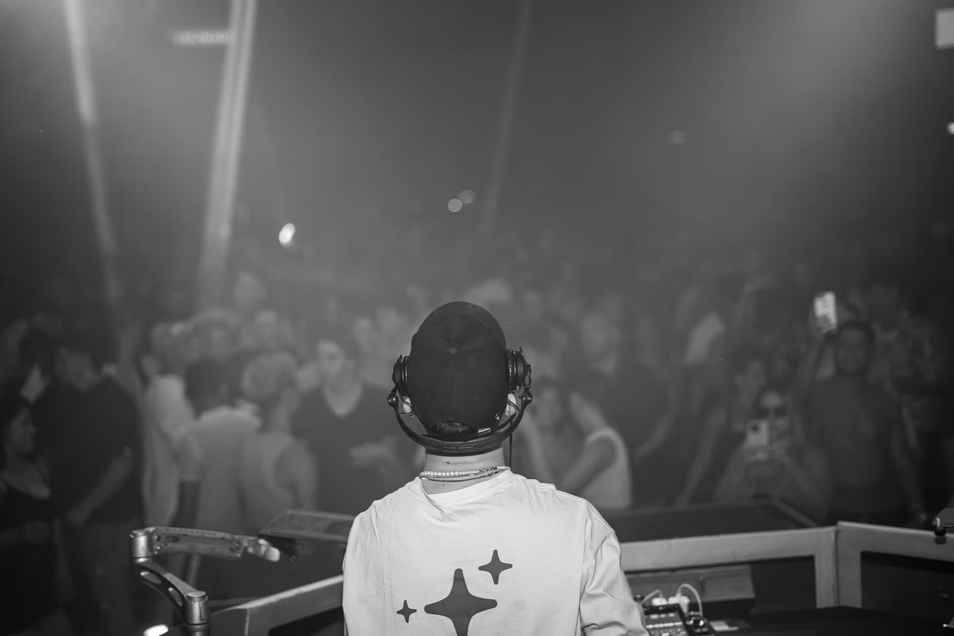 A DJ with headphones faces a large, blurry crowd in a dimly lit, smoky nightclub.