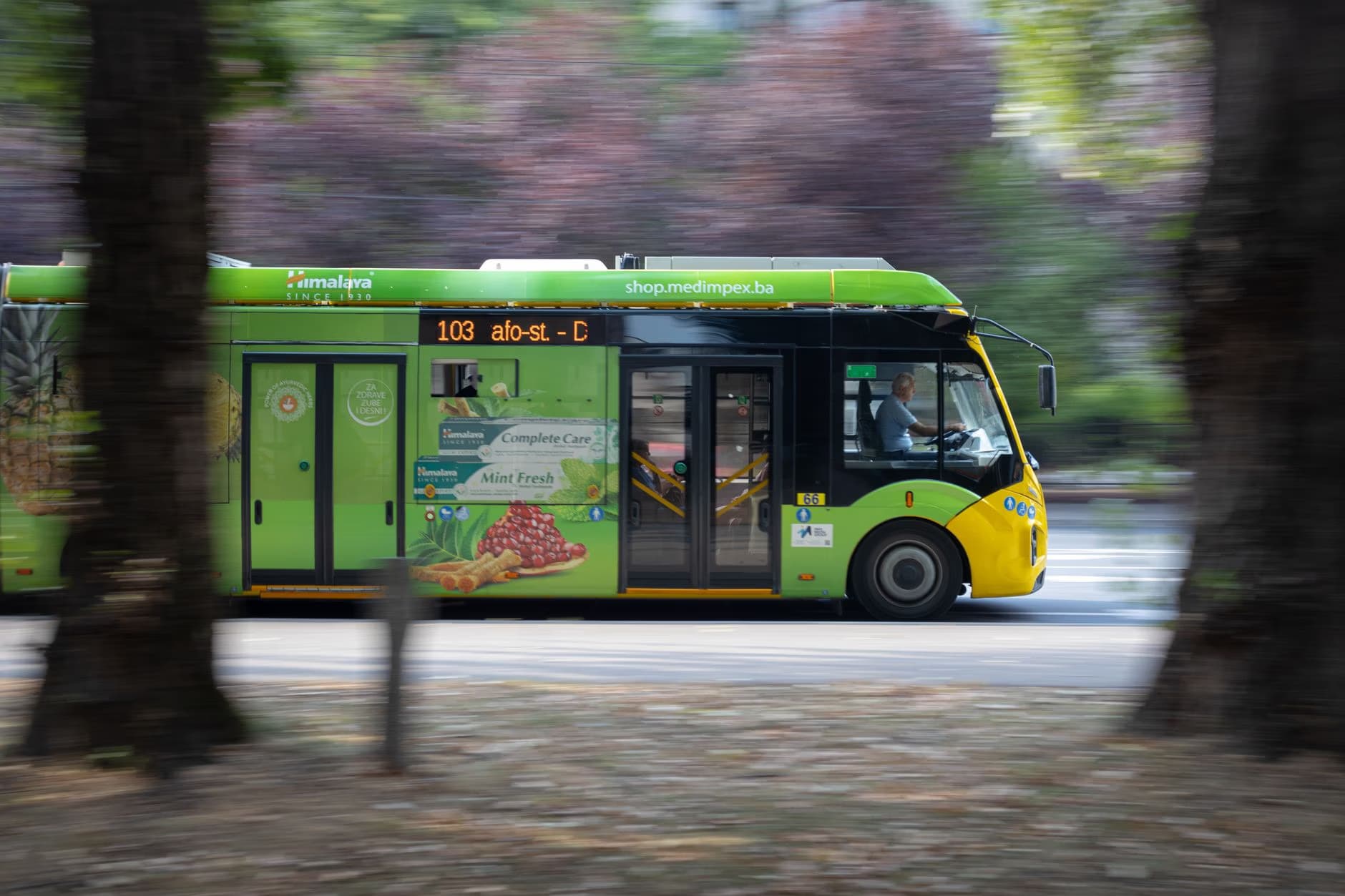A bright green city bus moves quickly past blurred trees along a street.