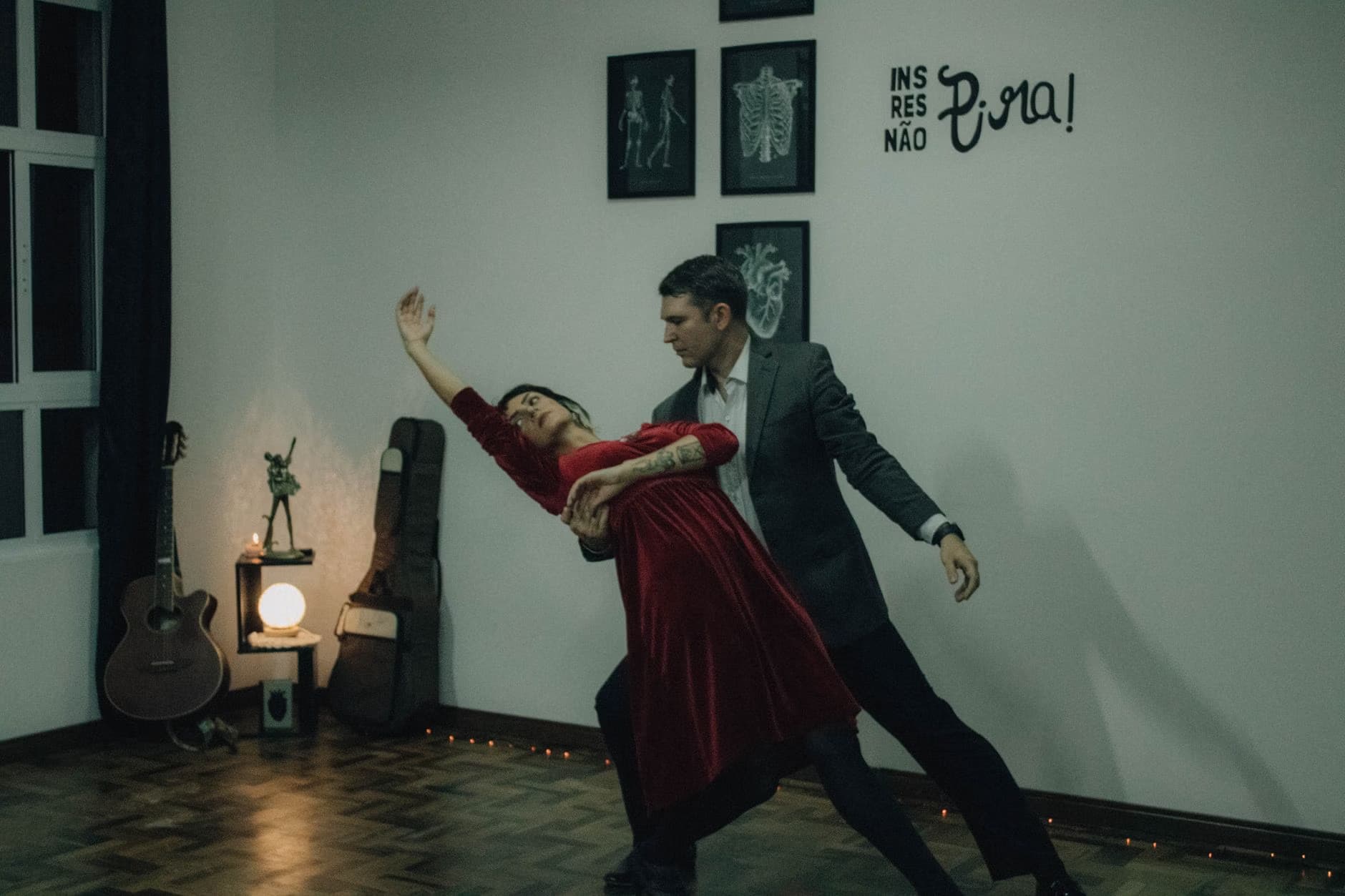 A man in a suit and a woman in a red dress perform a dance inside a dimly lit room.