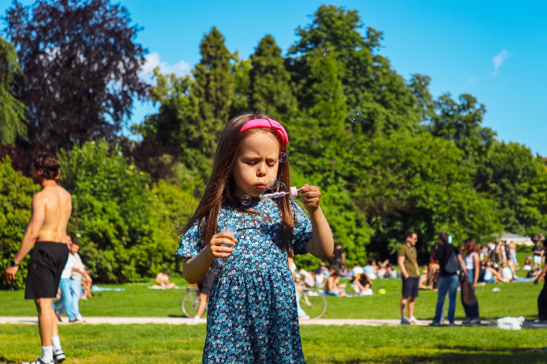 Mladá dívka v květovaných šatech fouká mýdlové bubliny ve slunném parku.