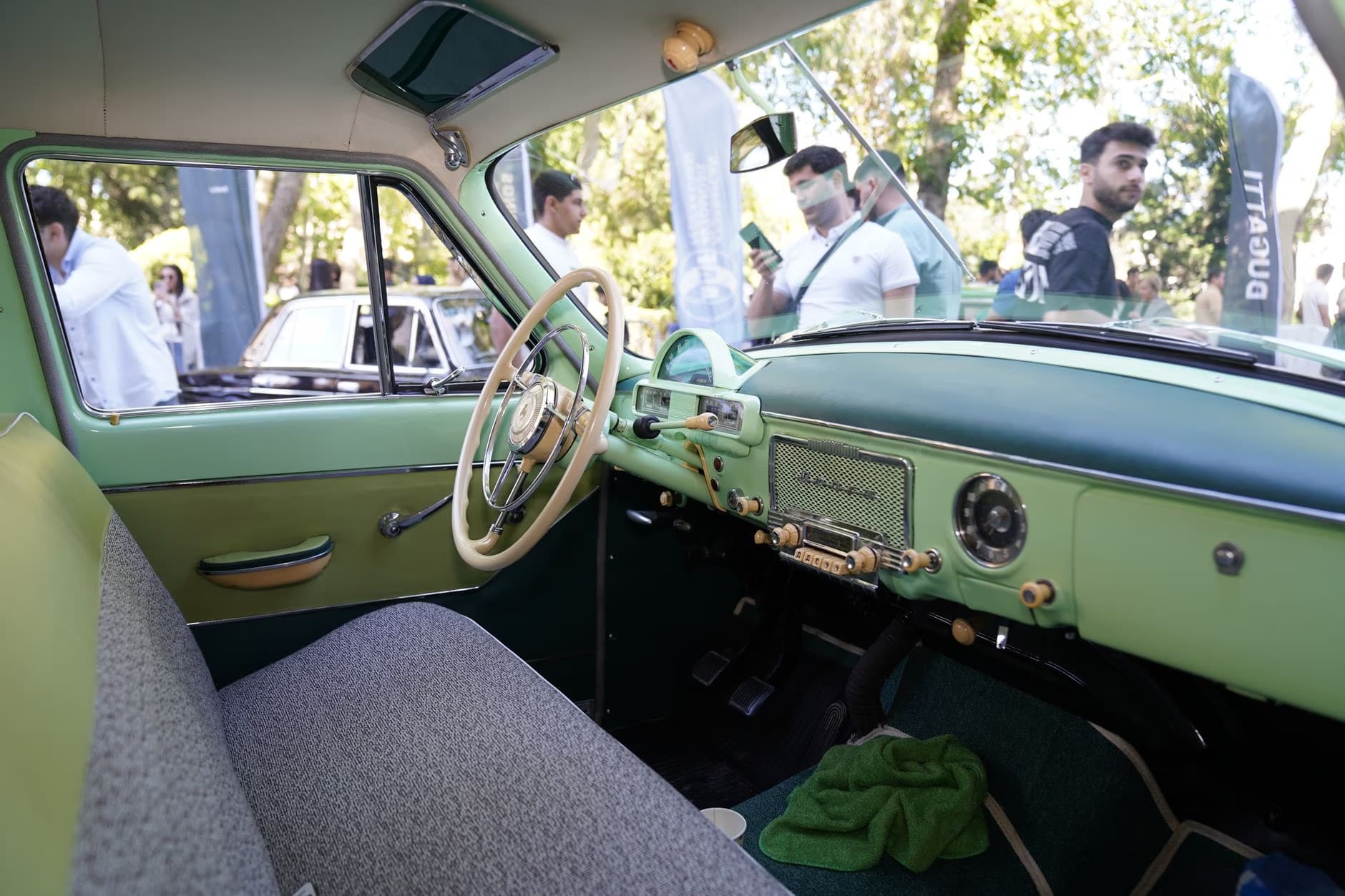 View from the light green interior of a vintage car at an outdoor event.