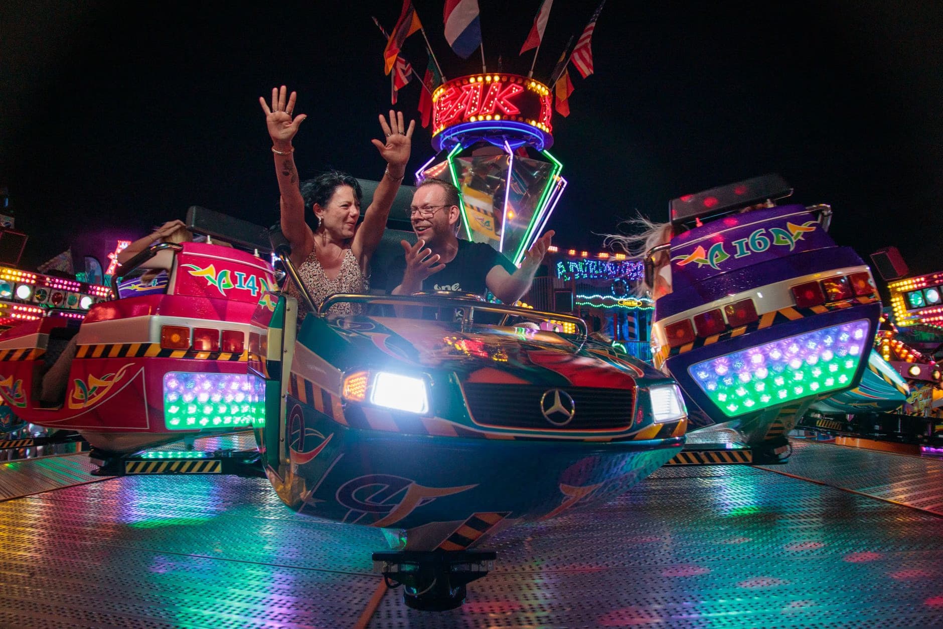 A laughing couple rides an illuminated fairground attraction with their hands raised at night.