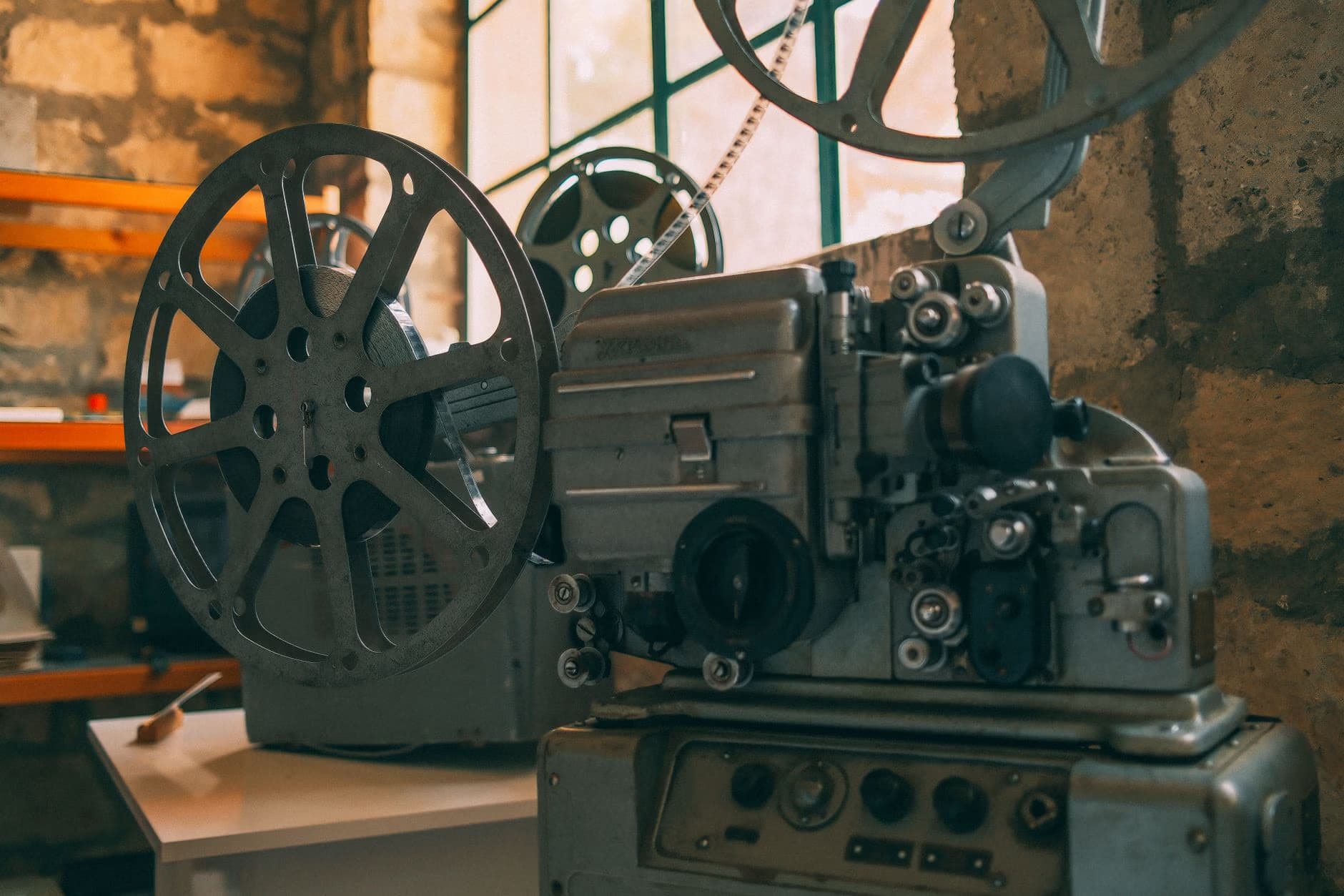 A vintage film projector with large metal reels sits on a table against stone walls.