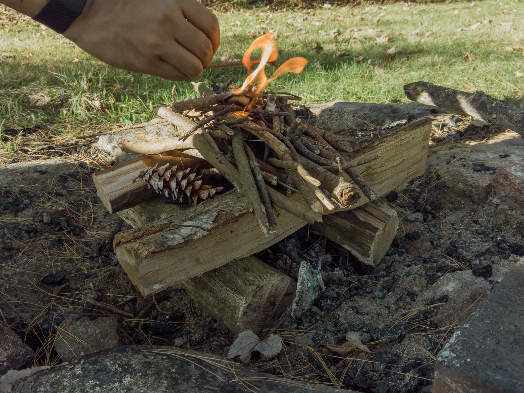 A hand places small twigs onto a small burning campfire in a rocky outdoor setting.