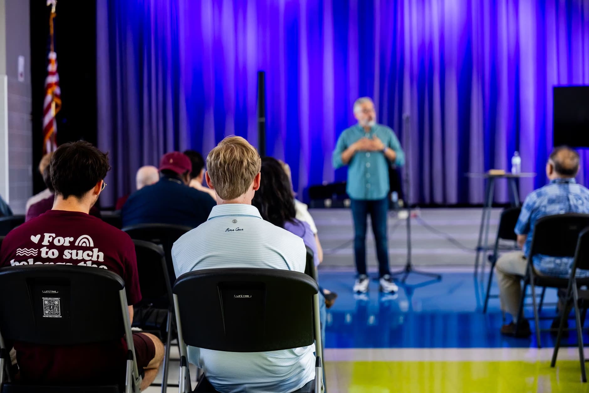 A man speaks on stage to an audience seated in chairs before purple curtains.