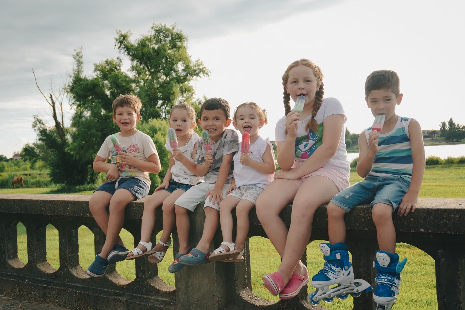 A group of six children sits on a low stone wall eating colorful ice pops outdoors.