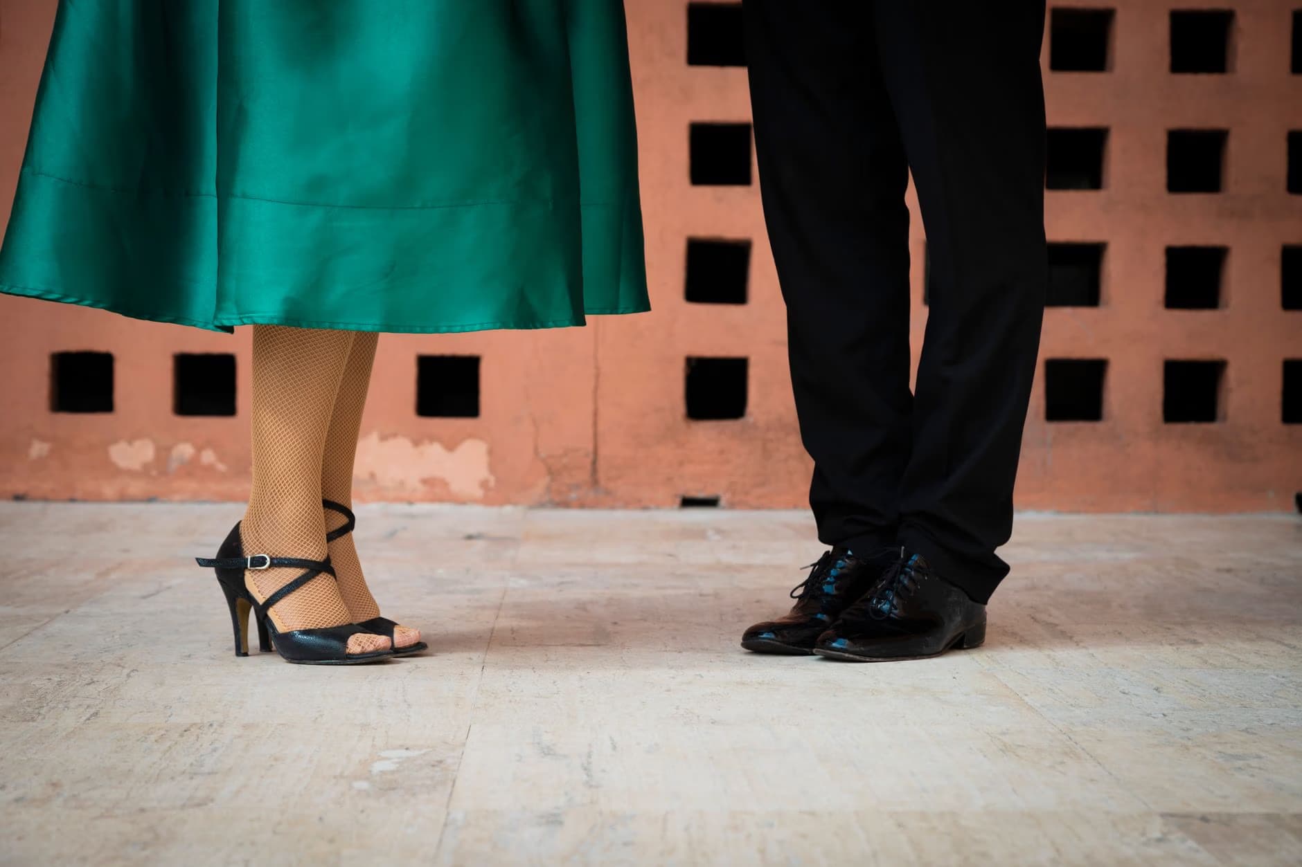 Lower bodies of a couple in formal wear standing on a stone floor before a patterned wall.