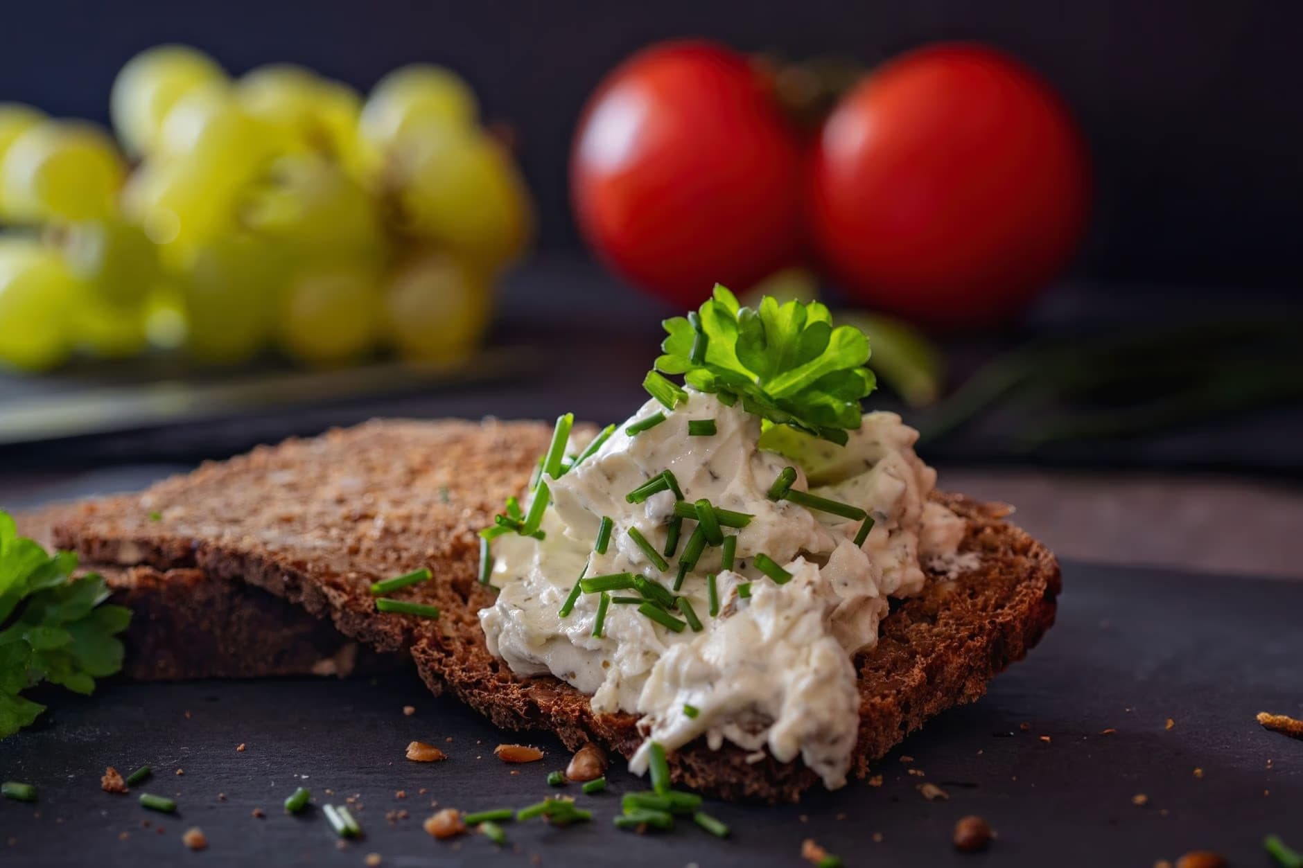 A slice of dark bread topped with creamy spread, fresh chives, and parsley on a dark surface.