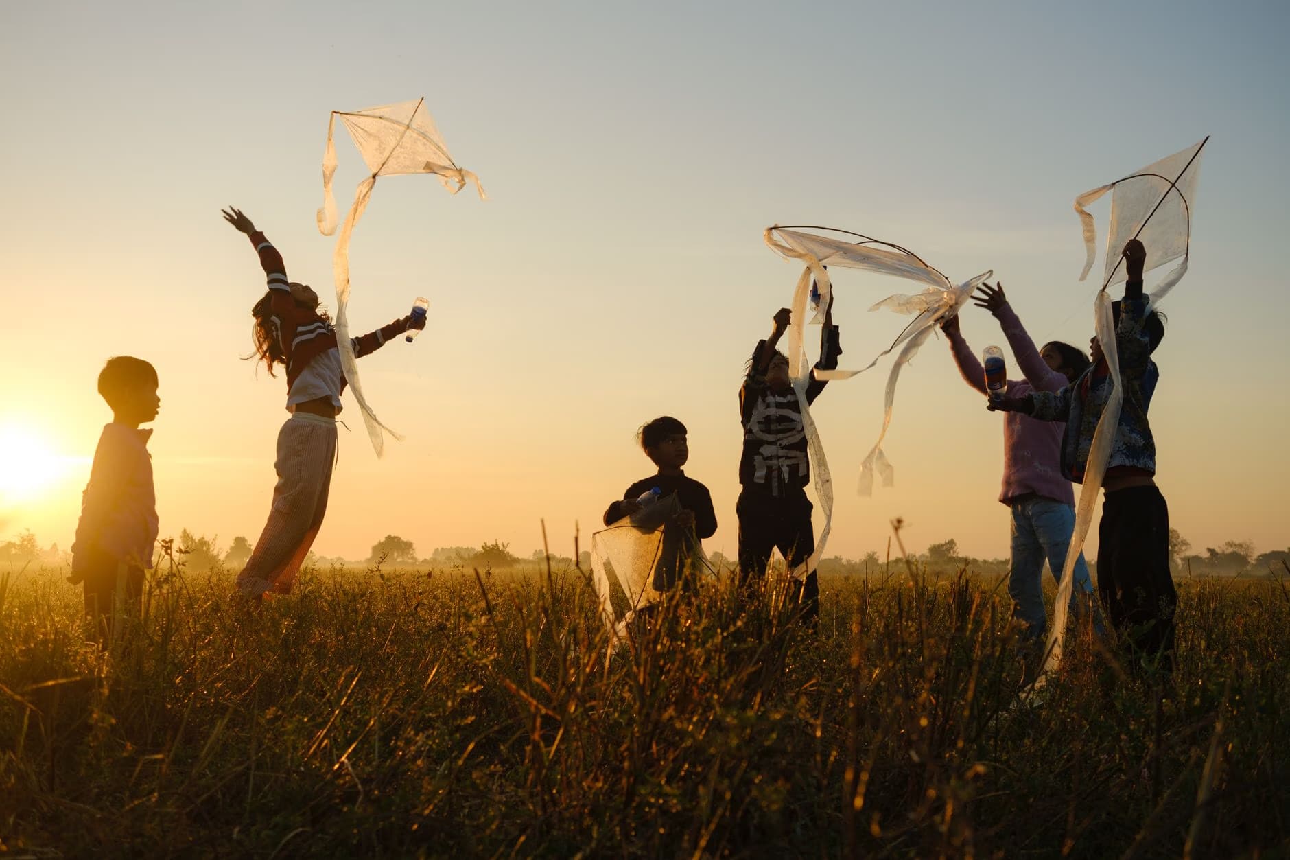 Silhouetted children run through a field while flying kites at sunset.
