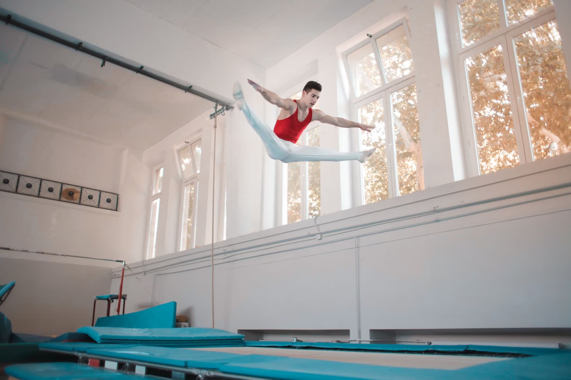 A male gymnast in a red leotard performs a high jump on a trampoline indoors.