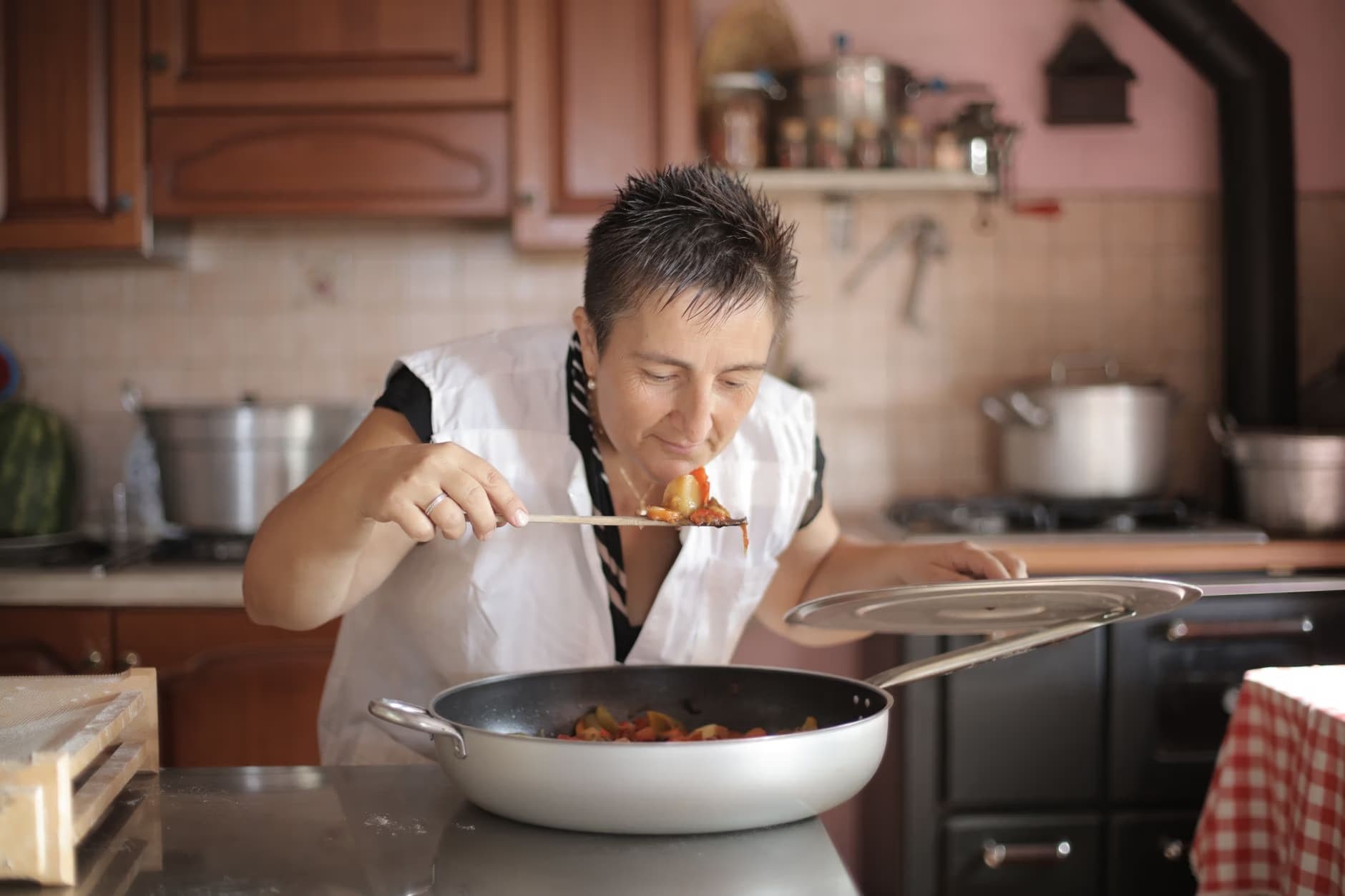A woman in a white apron tastes food with a wooden spoon in a kitchen.