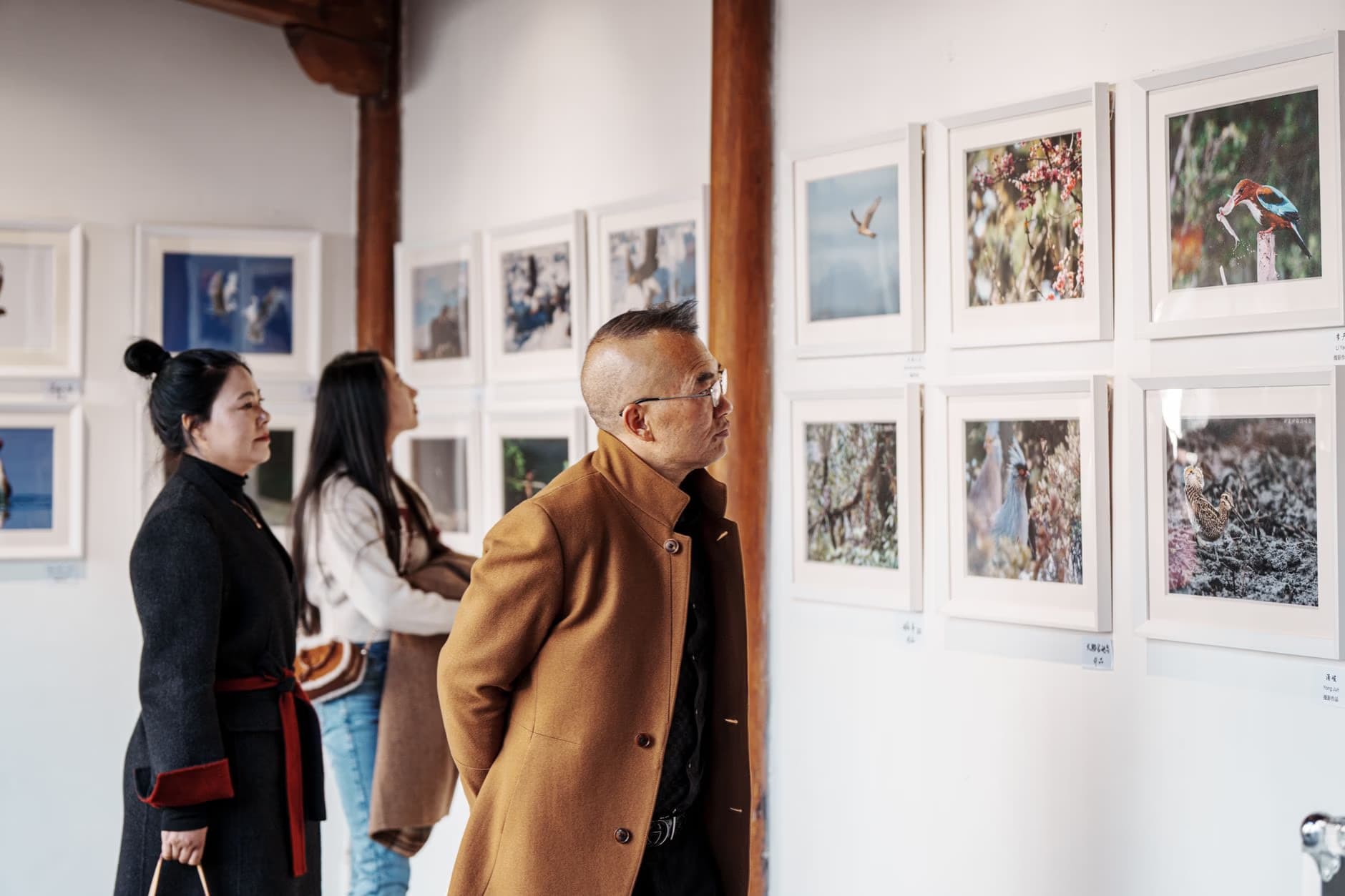 Three people observe framed artwork displayed on a white gallery wall.
