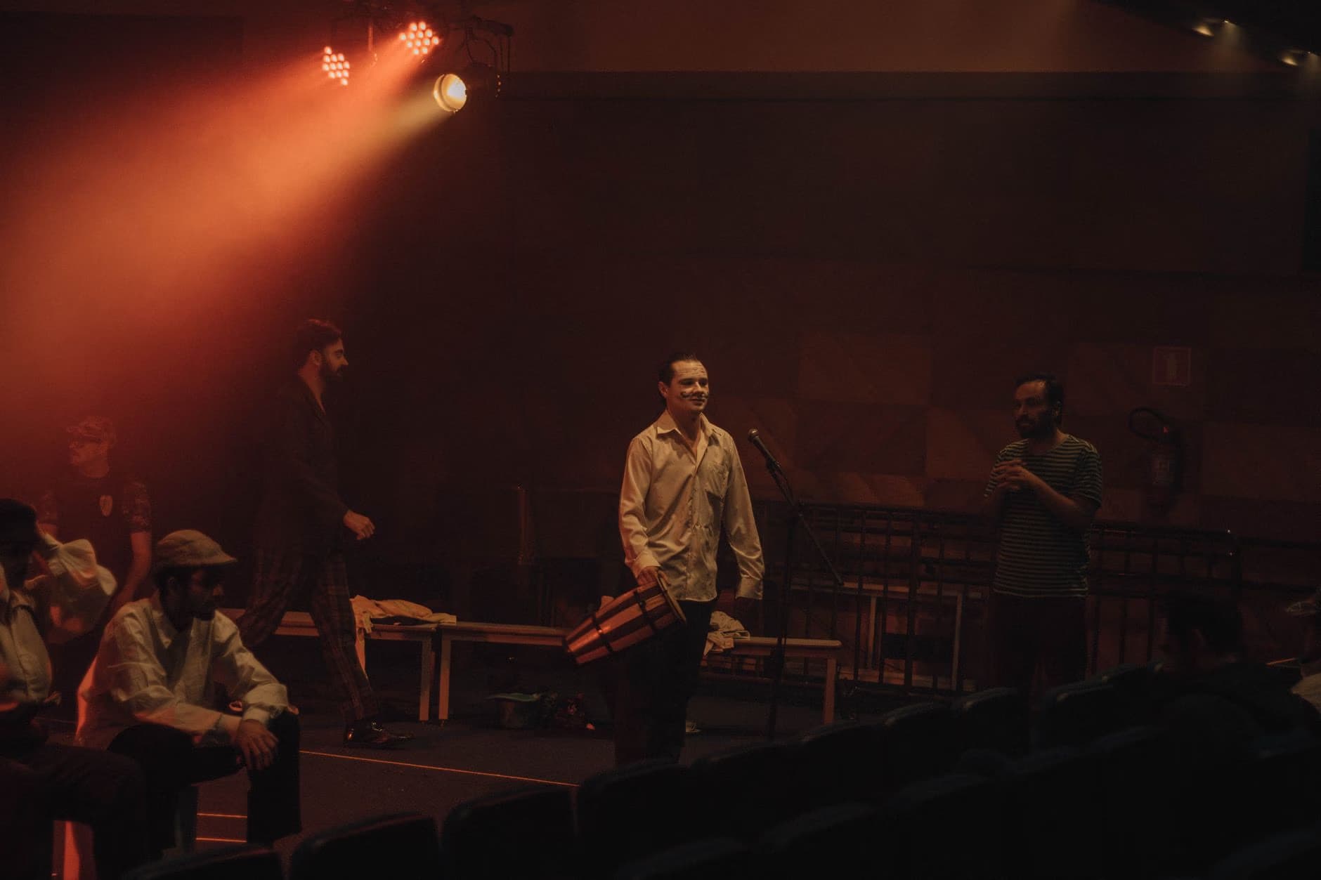 People stand on a dimly lit stage bathed in warm, reddish spotlight, carrying instrument cases.