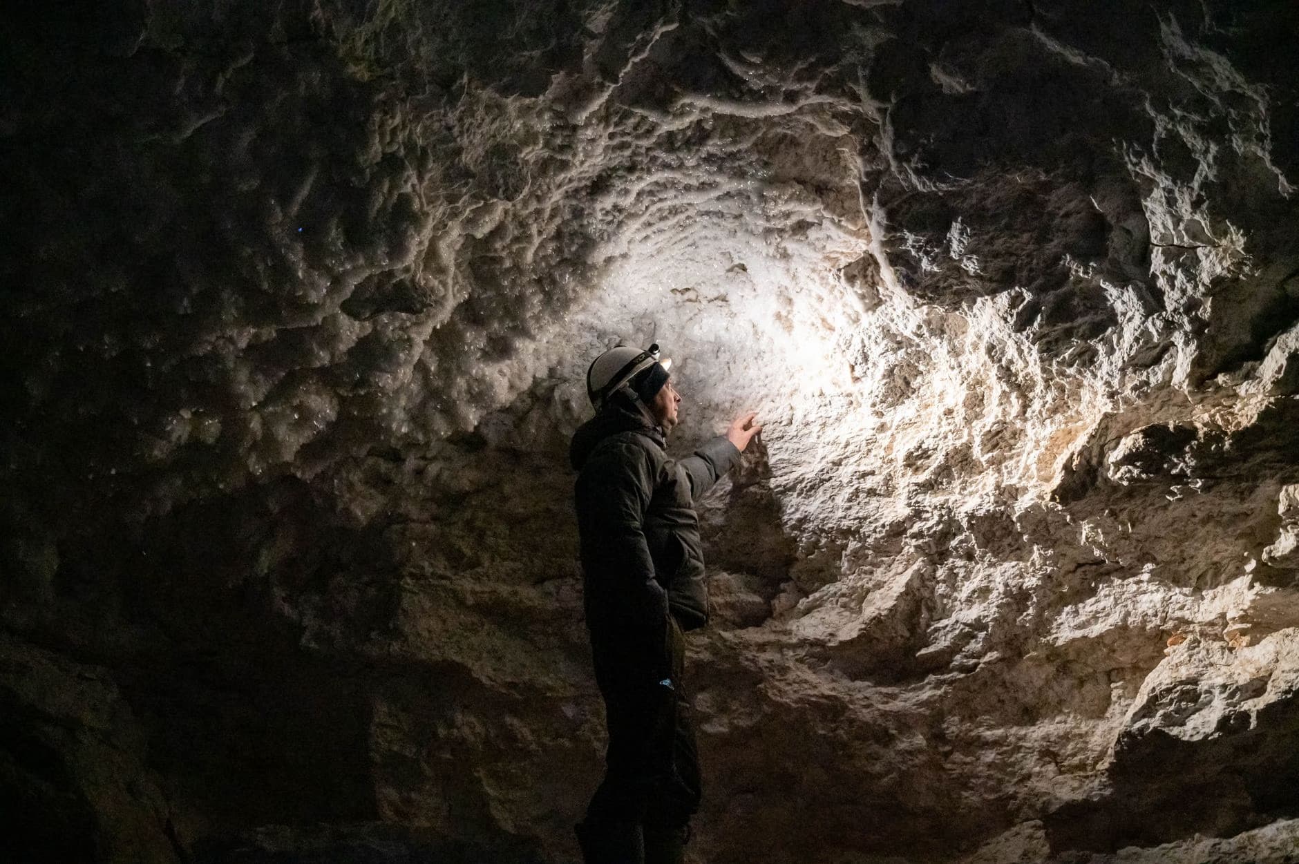 A person in a helmet explores a dimly lit cave tunnel, illuminating the rocky wall with a light.