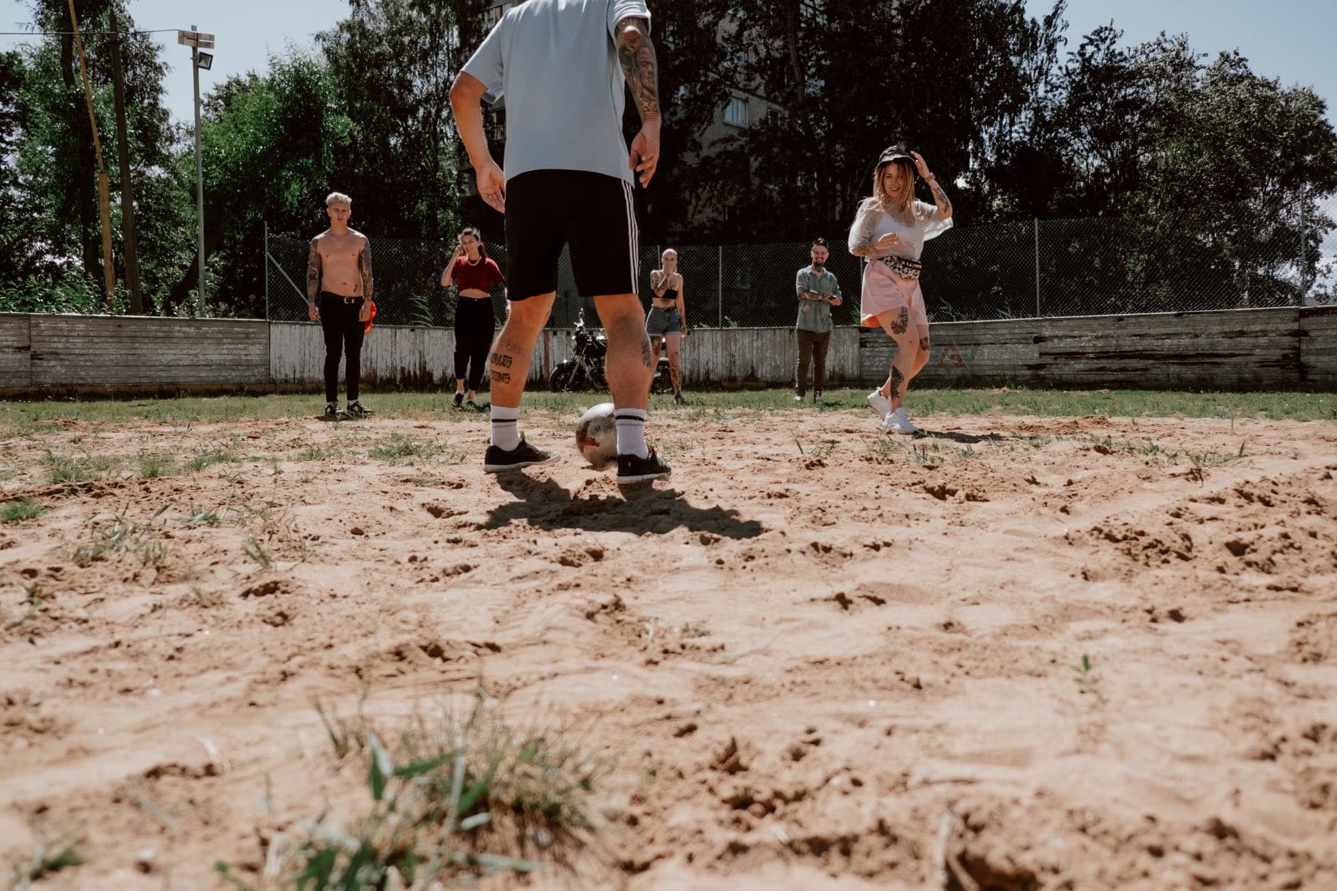 Skupina mladých lidí hraje za slunného dne venku fotbal na hřišti s hlinitým povrchem.