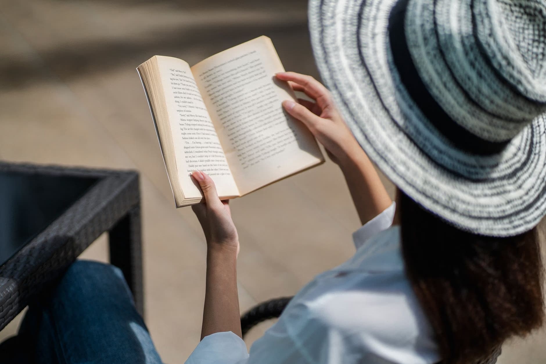 A woman wearing a sun hat holds an open book while sitting outside.