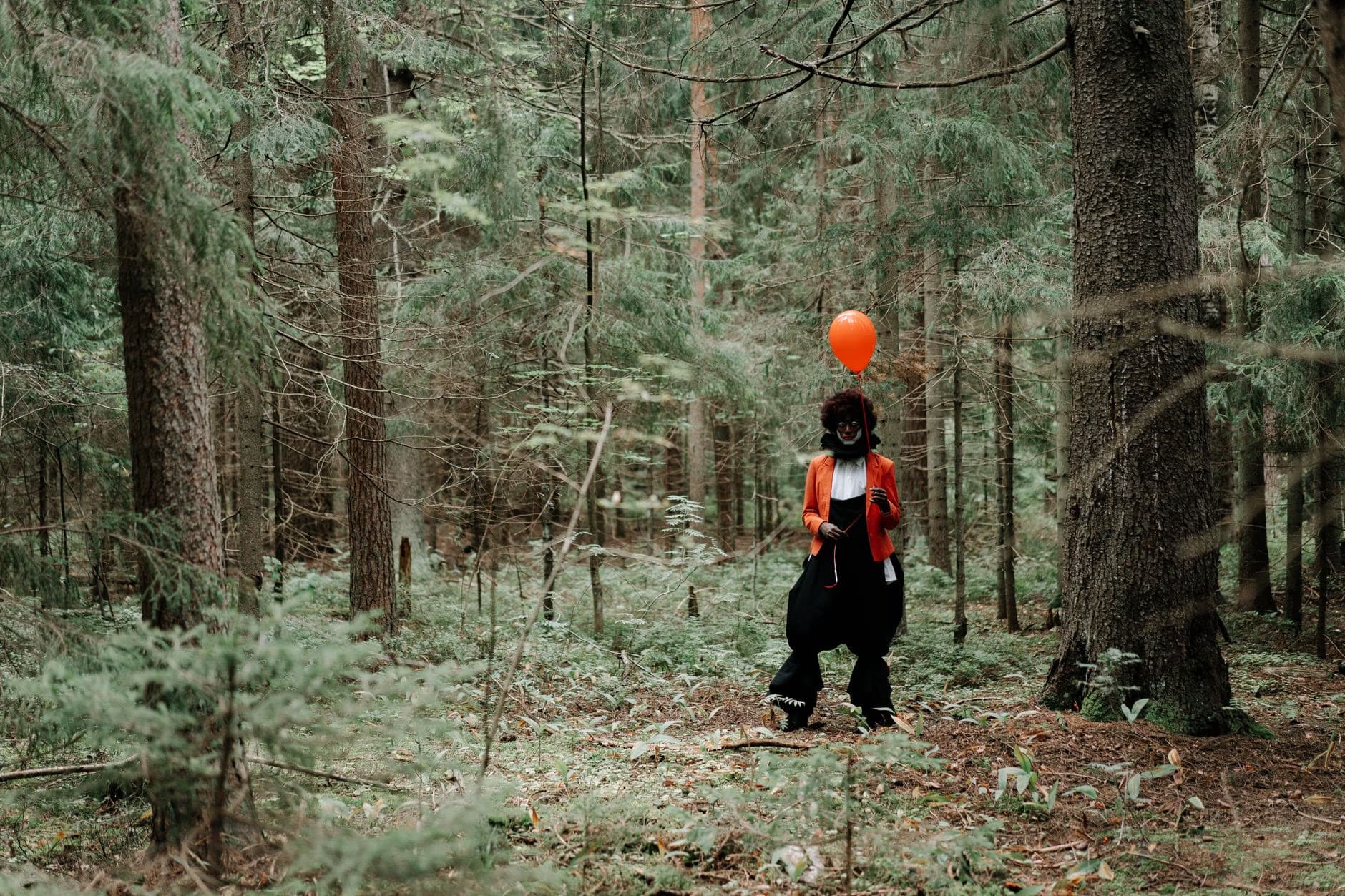 A clown in an orange jacket holds a balloon while standing in a dense coniferous forest.