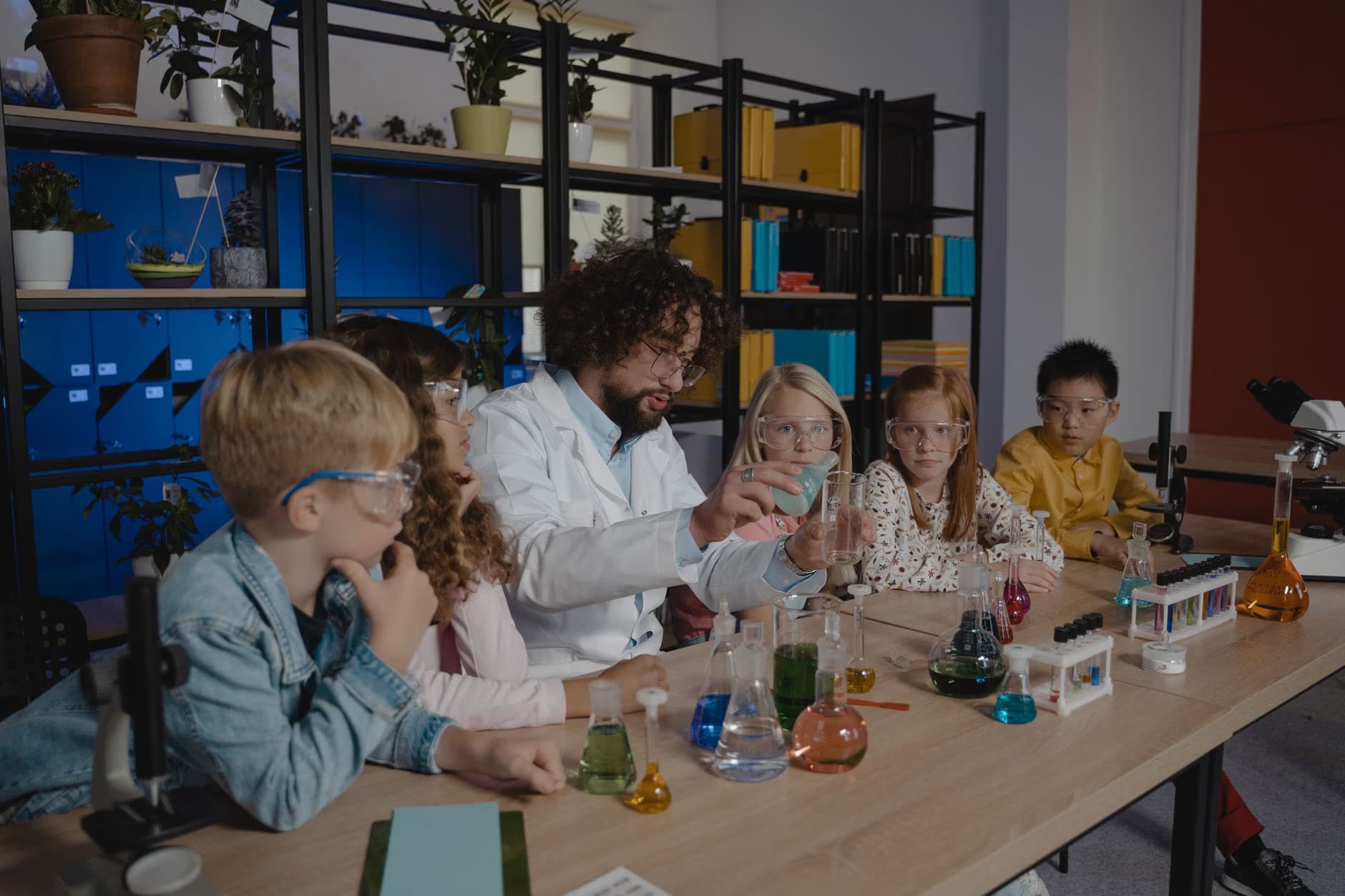 Učitel v laboratorním plášti předvádí dětem ve třídě chemický pokus.