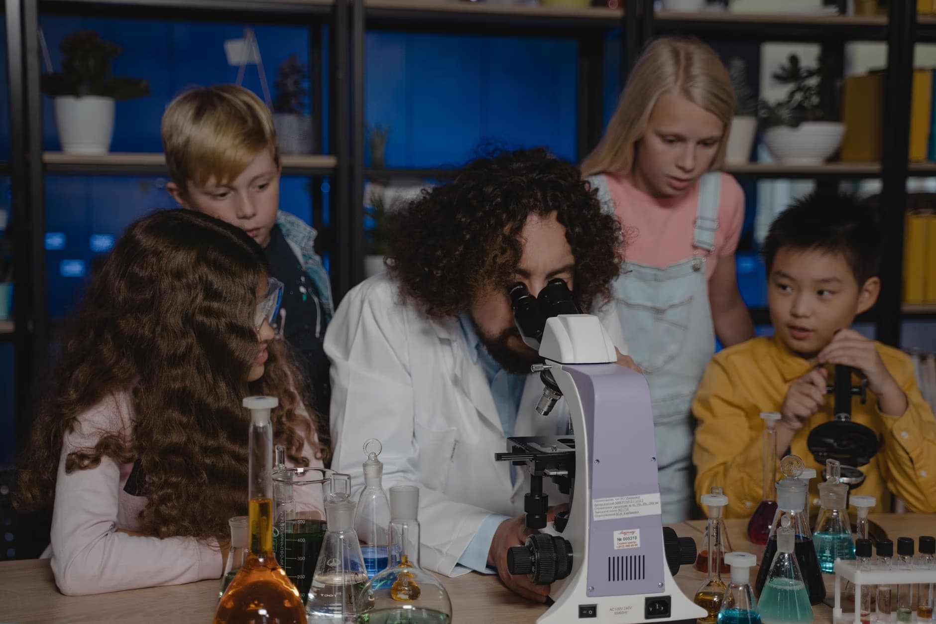 Učitel v laboratorním plášti ukazuje dětem ve třídě, jak pracovat s mikroskopem.