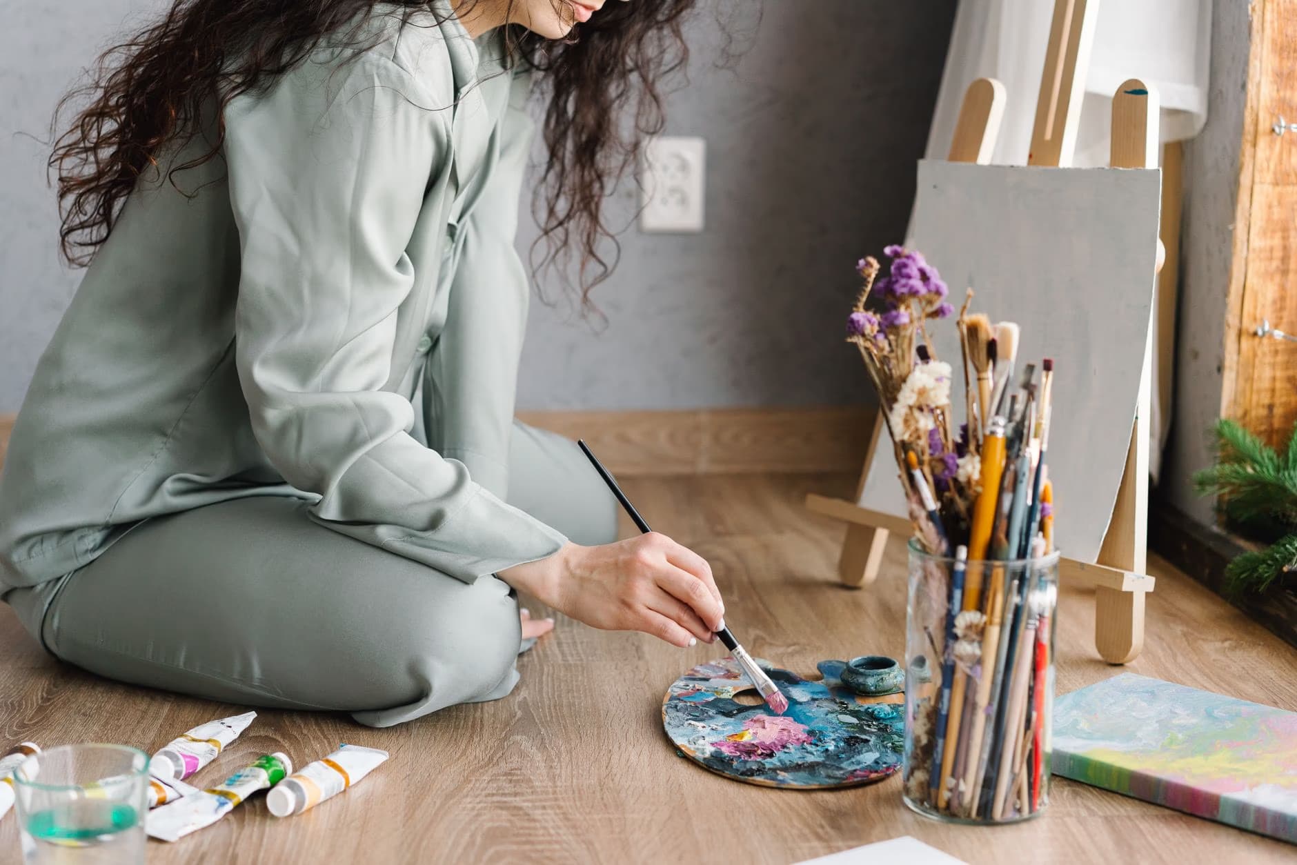 A woman with curly hair sits on the floor painting on a palette with a brush.
