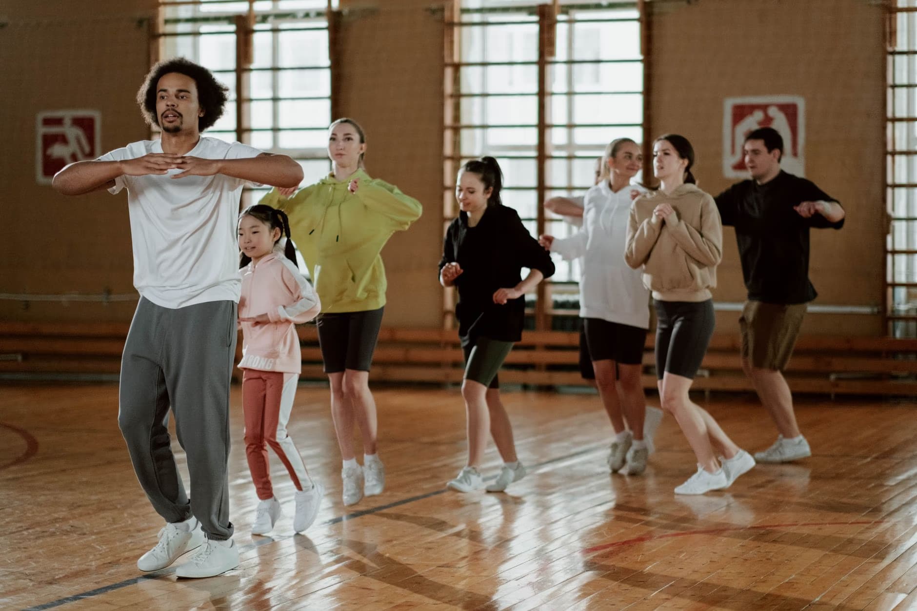 A diverse group of people follows an instructor in a dance or exercise class inside a gym.