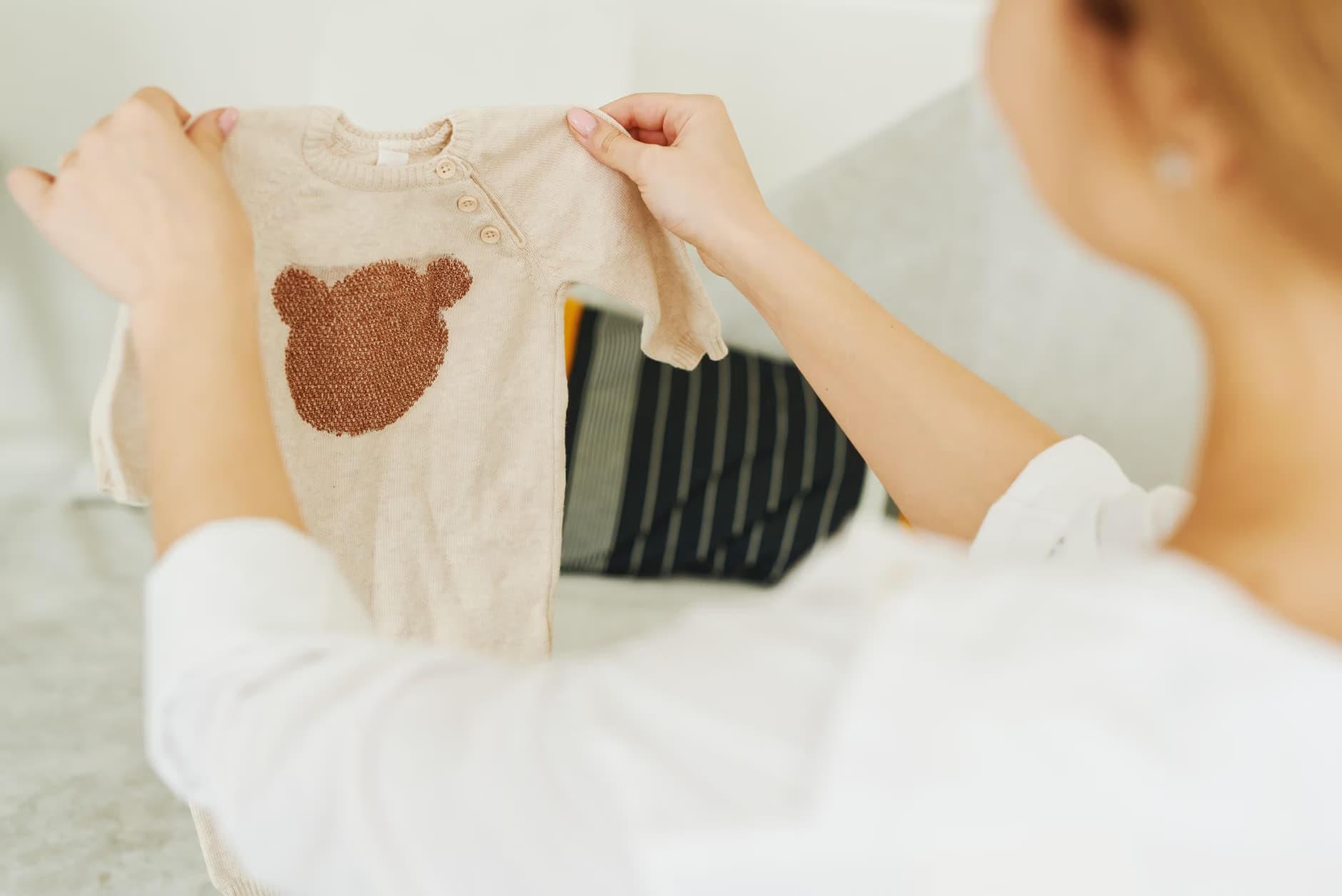 Person holds up a beige baby onesie decorated with a brown teddy bear patch.
