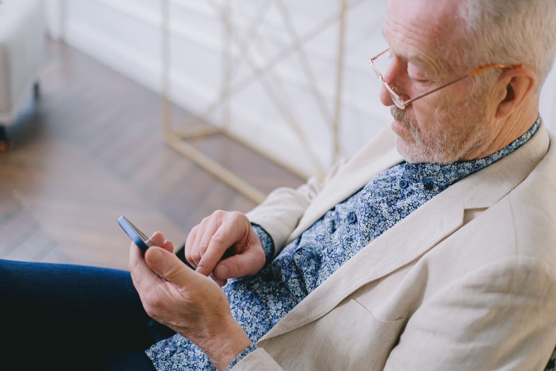 Older man in beige blazer and glasses looks at his smartphone while seated indoors.