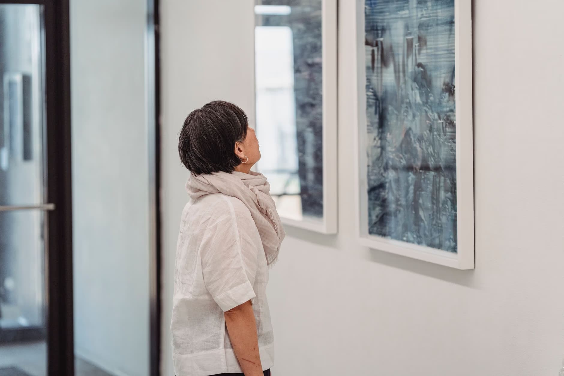 A woman with a scarf observes abstract paintings displayed on a gallery wall.