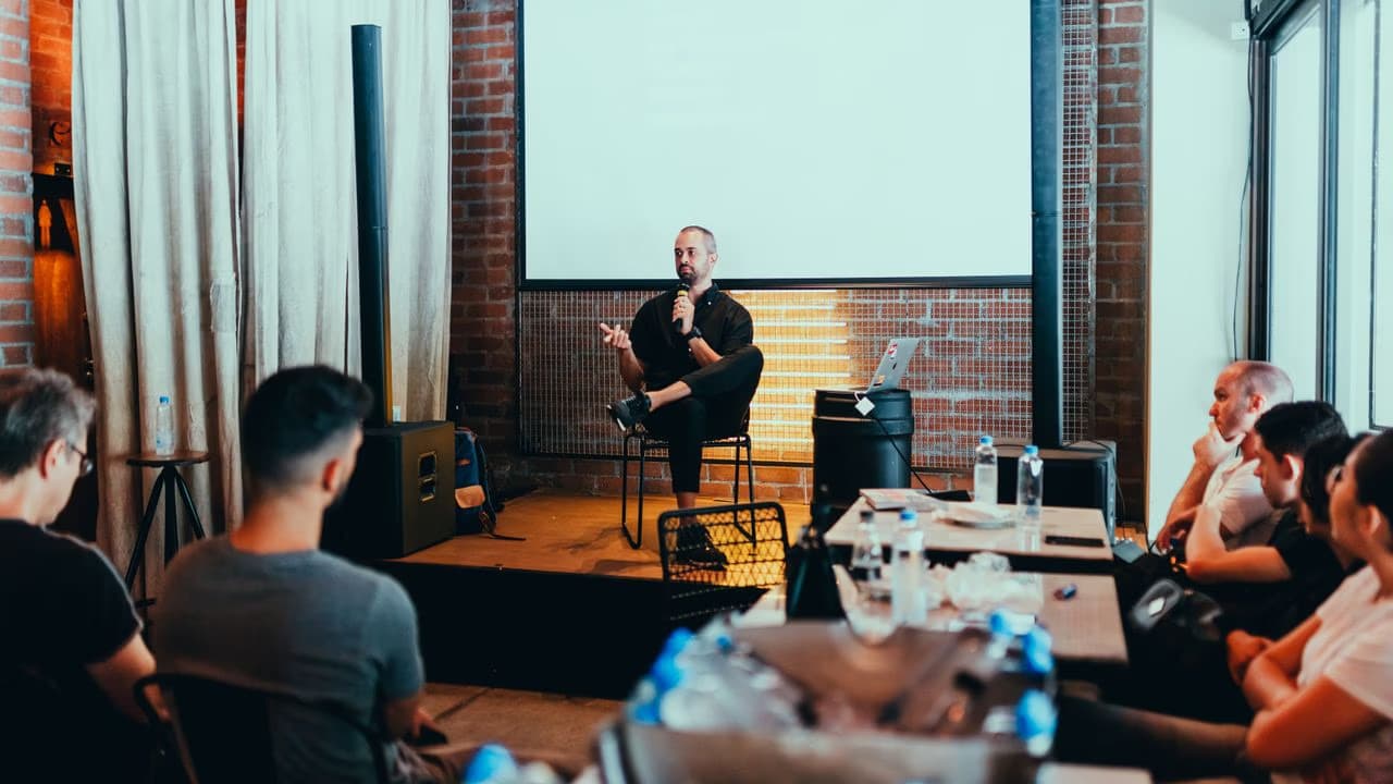A man sits on a stage speaking to an audience in a brick-walled room.