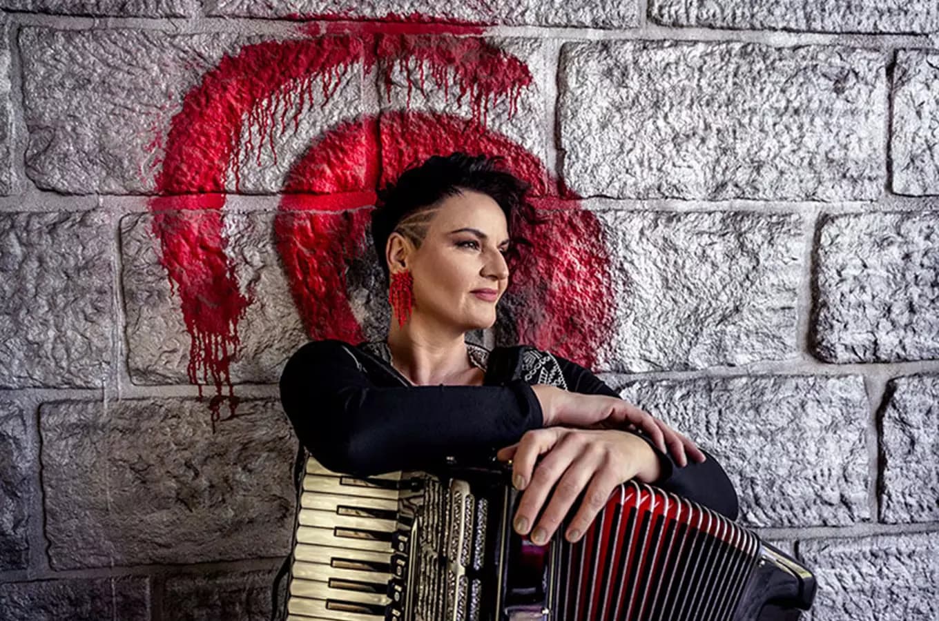 Žena s krátkými tmavými vlasy pózuje s akordeonem před kamennou zdí s červenými malovanými ornamenty.
