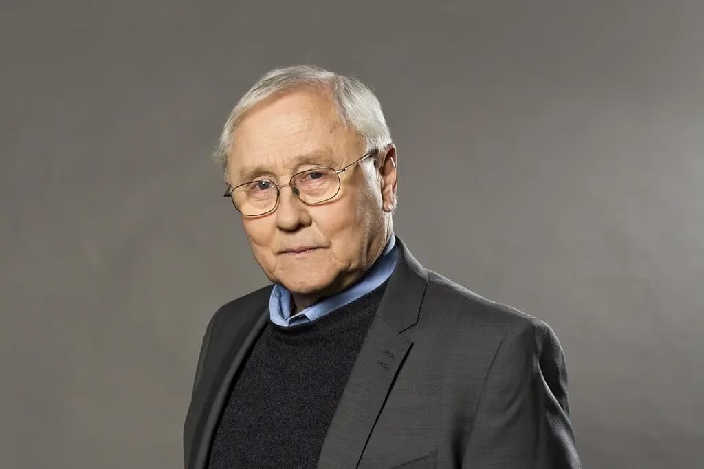 Older man with glasses wearing a dark jacket over a light blue shirt against grey.
