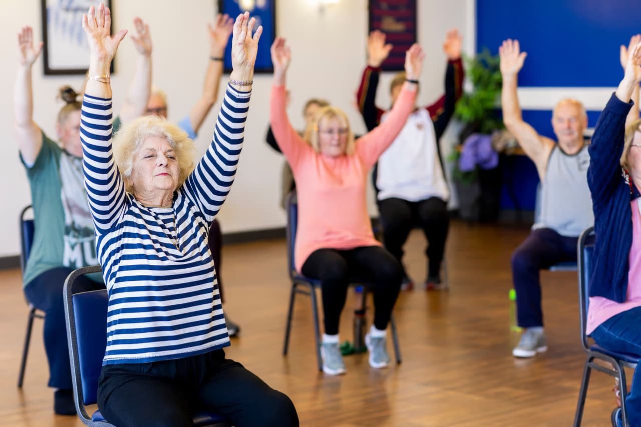 A group of older adults sit on chairs in a room exercising with arms raised overhead.
