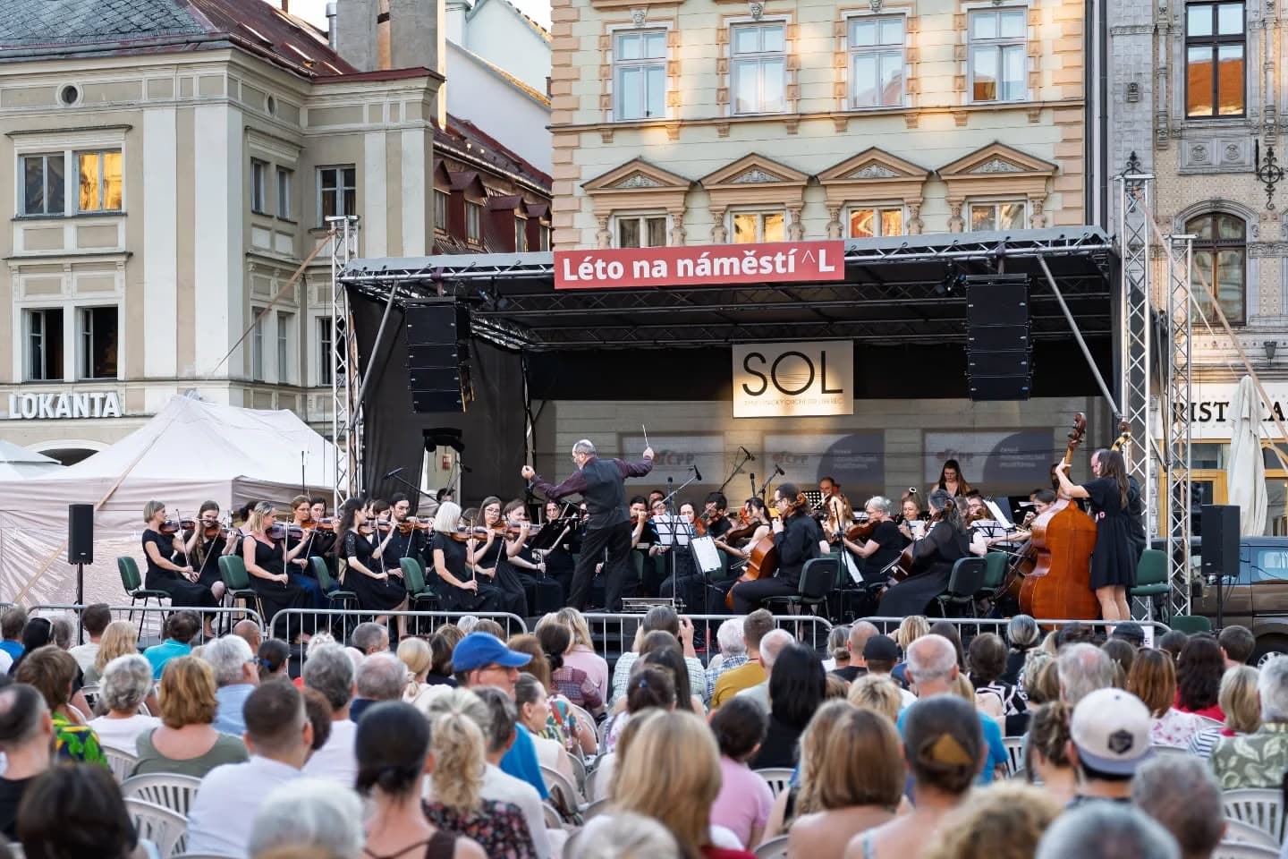 Ein Orchester tritt auf einer Außenbühne auf einem Stadtplatz vor einem großen Publikum auf.
