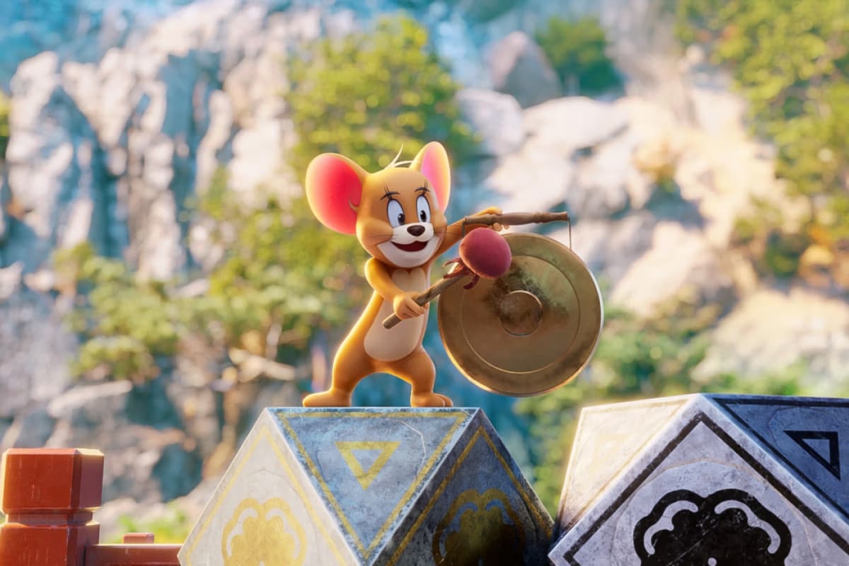 Jerry the mouse hits a brass gong with a mallet, standing before a mountainous landscape.