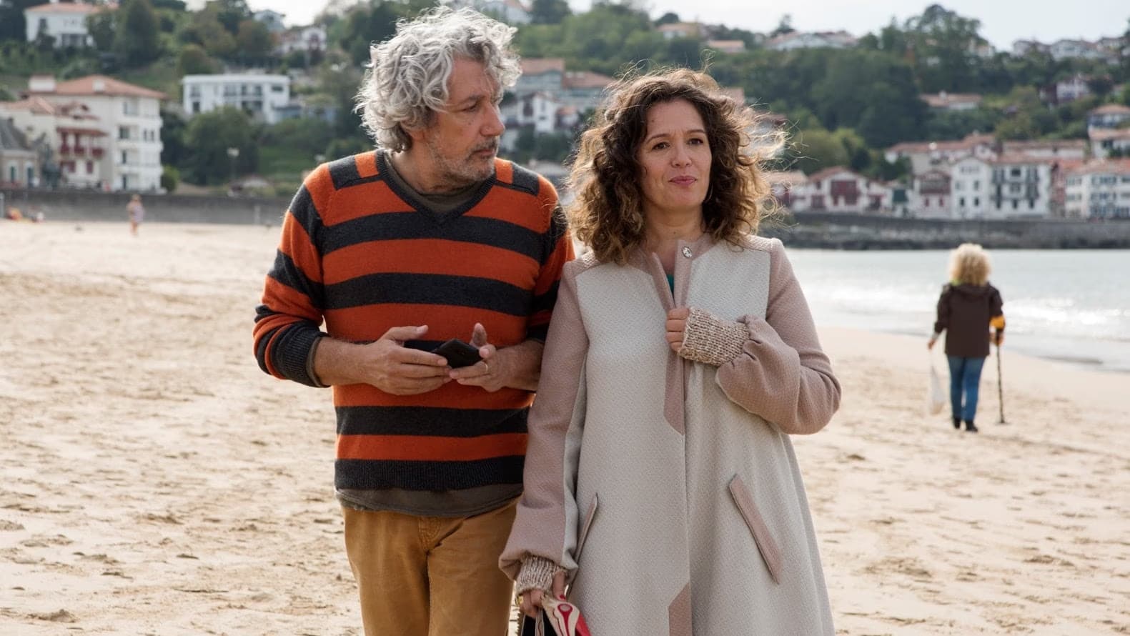 A middle-aged man and woman walk together on a sandy beach by the sea.