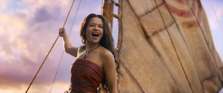 A young woman stands joyfully on a sailboat at sea during sunset.