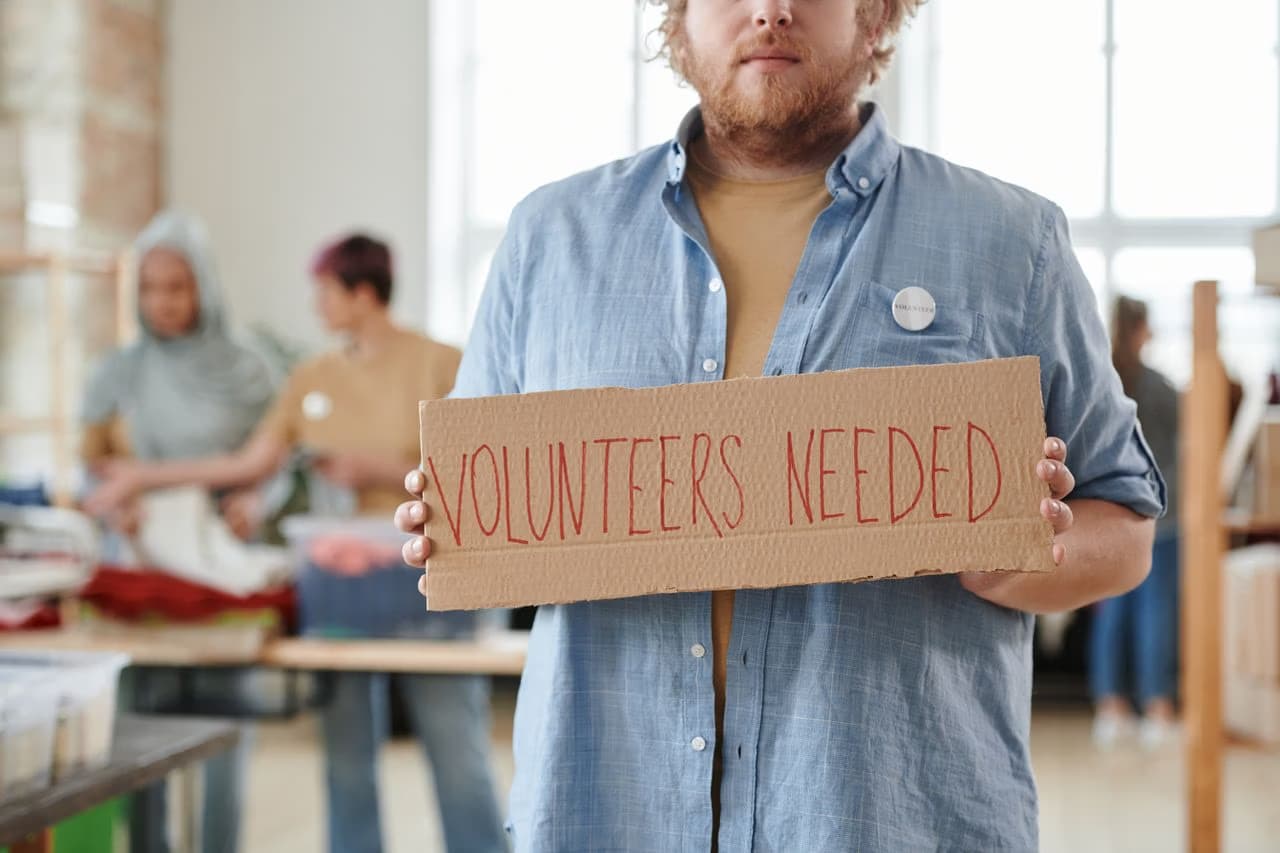 Muž drží v ruce kartonovou ceduli s nápisem Volunteers Needed v prostorách skladu.