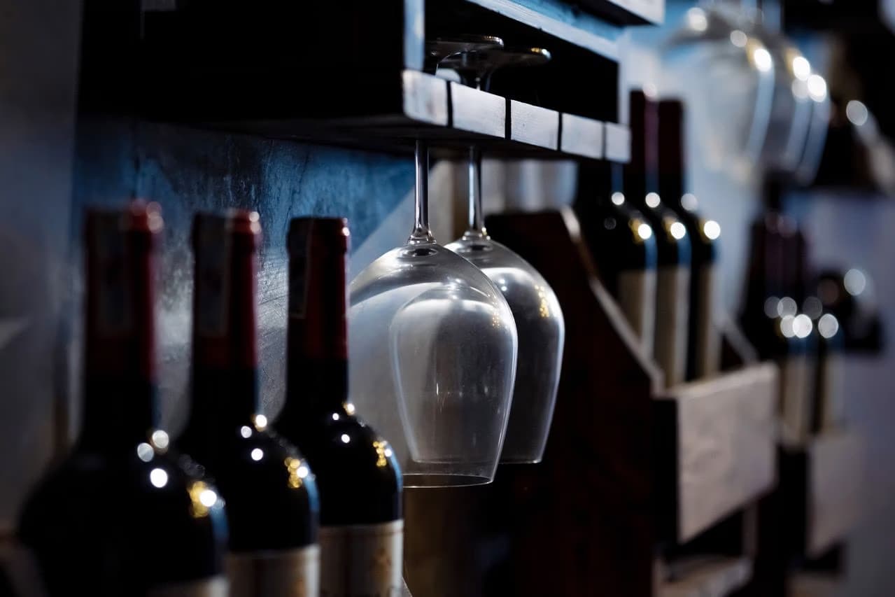Wine bottles and hanging stemmed glasses on a rustic wooden rack in dim light.