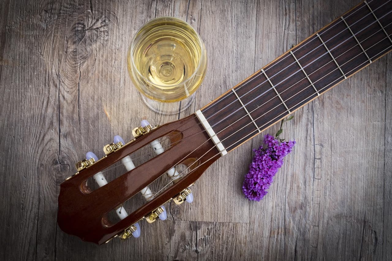 Guitar neck, a glass of white wine, and purple lilac flowers rest on a wooden table.