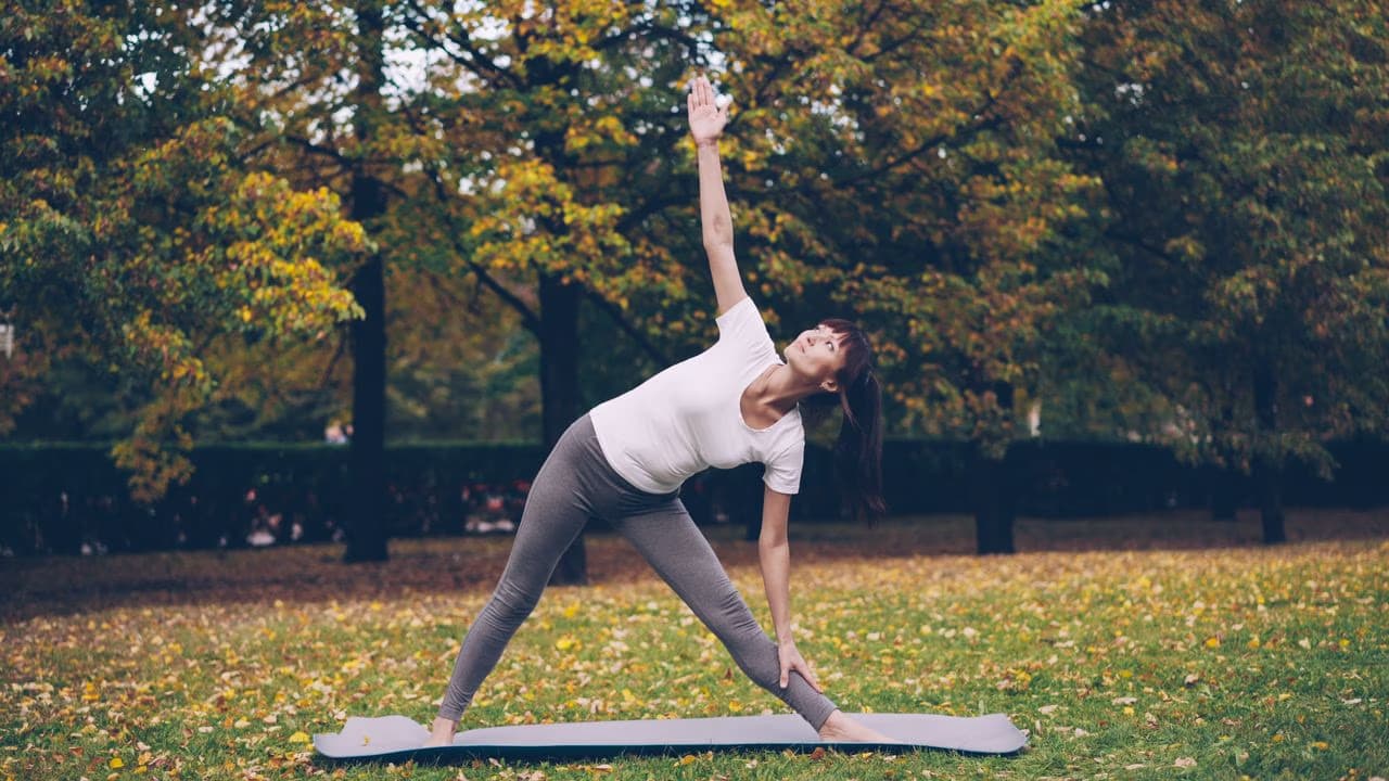 A woman performs a yoga triangle pose on a mat in an autumnal park setting.